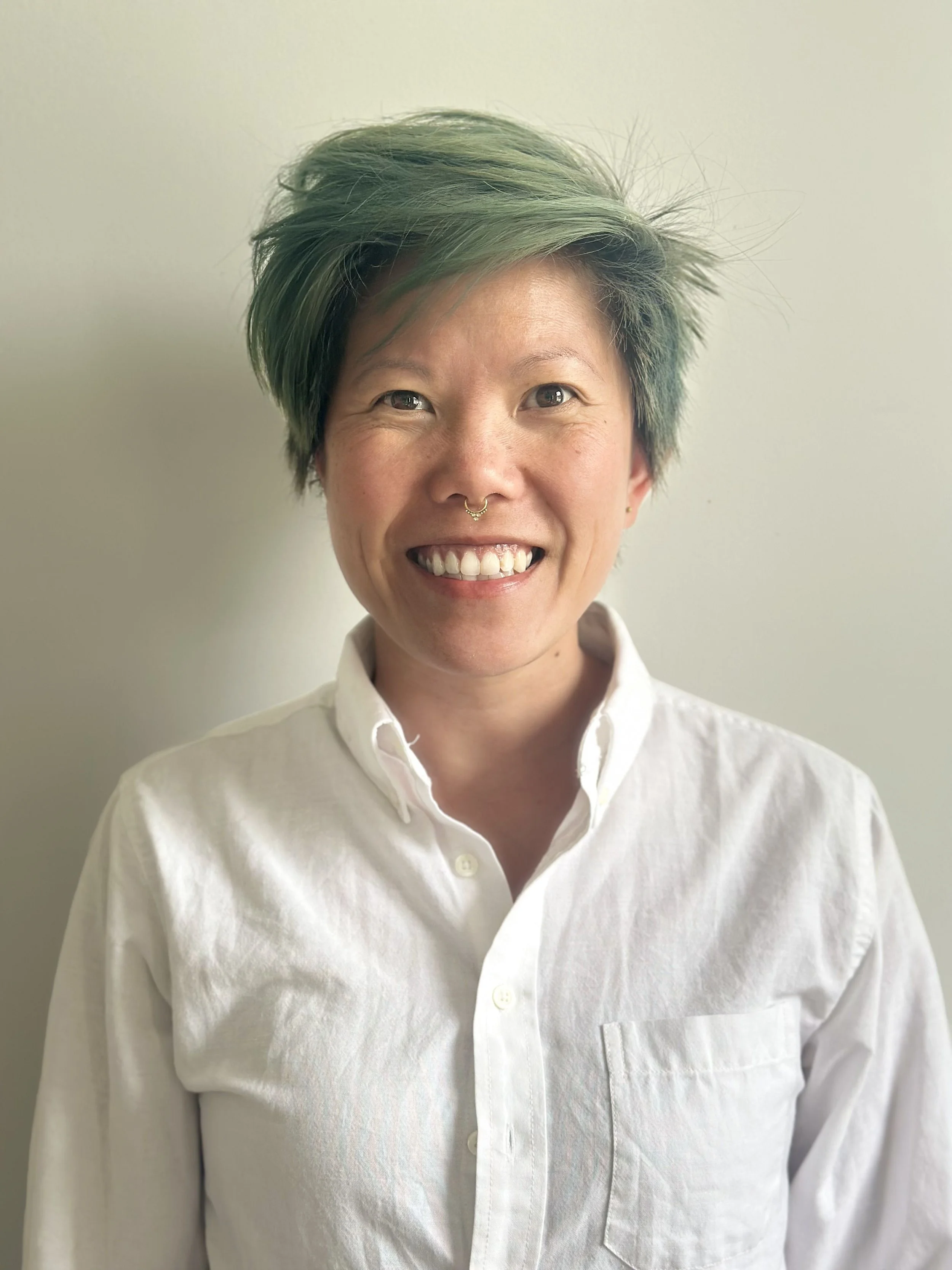 Person with short green hair smiling, wearing a white button-up shirt, against a plain light-colored background.