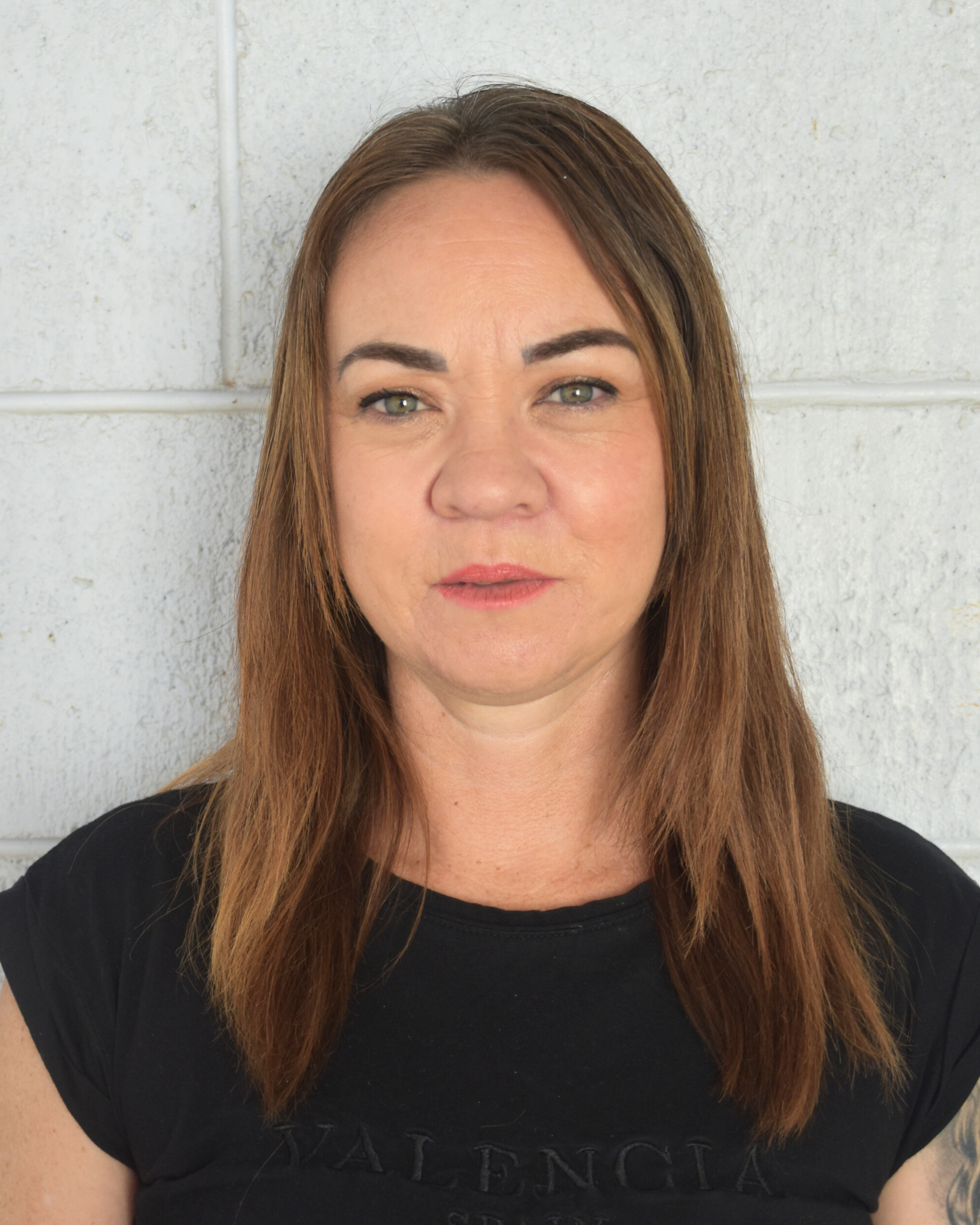 Close-up of a woman with wavy brown hair and a tattoo on her right shoulder, smiling against a white brick wall background.