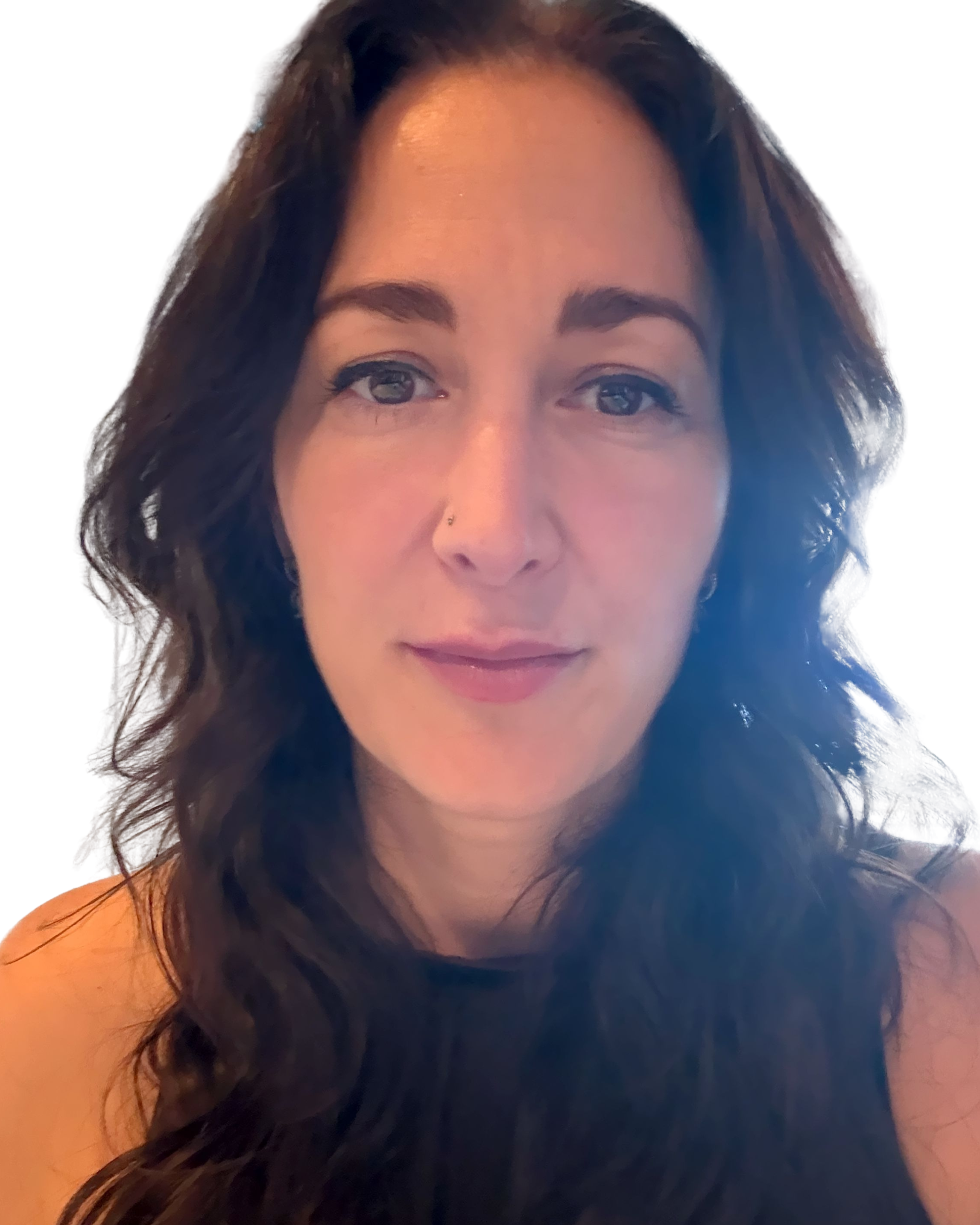 Close-up portrait of a woman with long, wavy dark hair, light makeup, and a small nose piercing, wearing a black top against a plain white background.