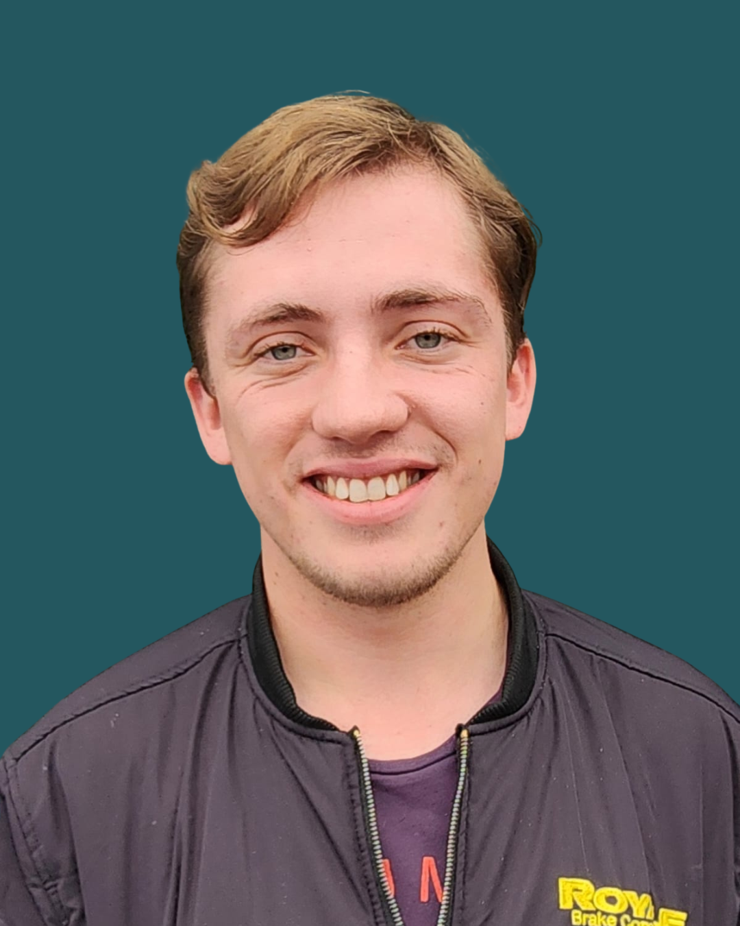 A young man with light brown hair smiling, wearing a black jacket with a yellow and red logo, against a dark teal background.