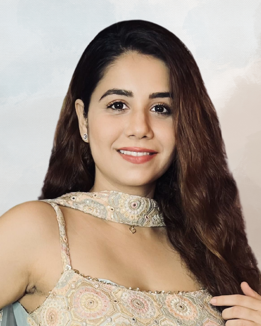 A woman with long brown hair smiling, wearing a detailed pastel-colored dress with embroidery and a sheer neckline, and small earrings.