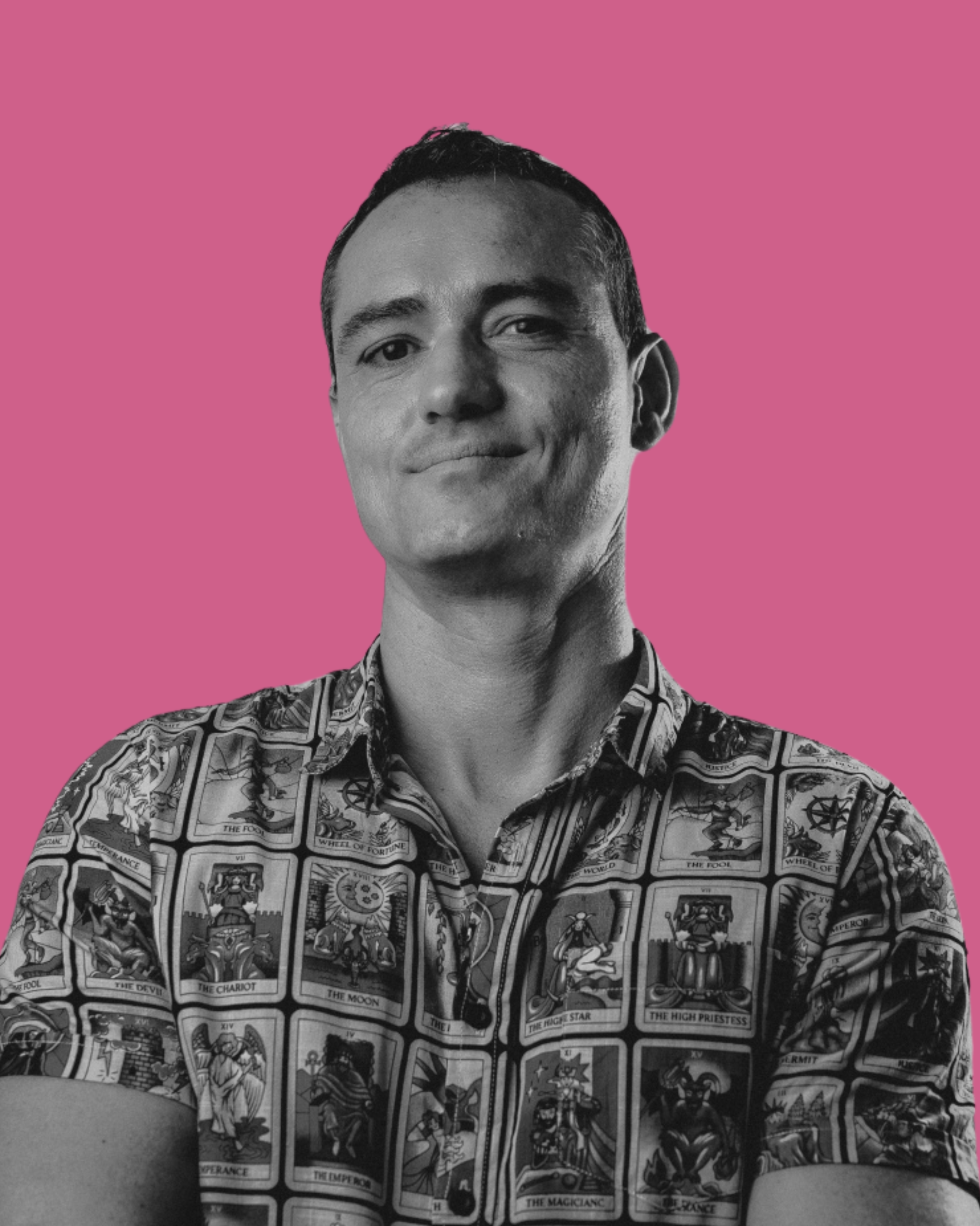 A black and white photo of a man in a short-sleeve button-up shirt with tarot card designs, standing against a pink background, smiling slightly.