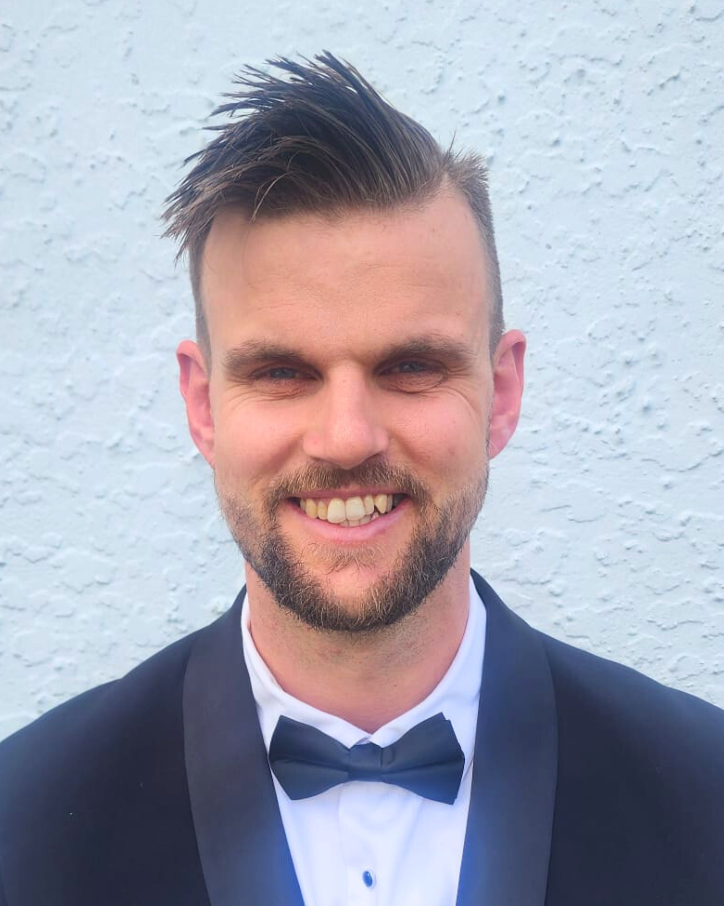 A smiling man with dark hair and a beard, wearing a black tuxedo and bow tie, standing against a white textured wall background.