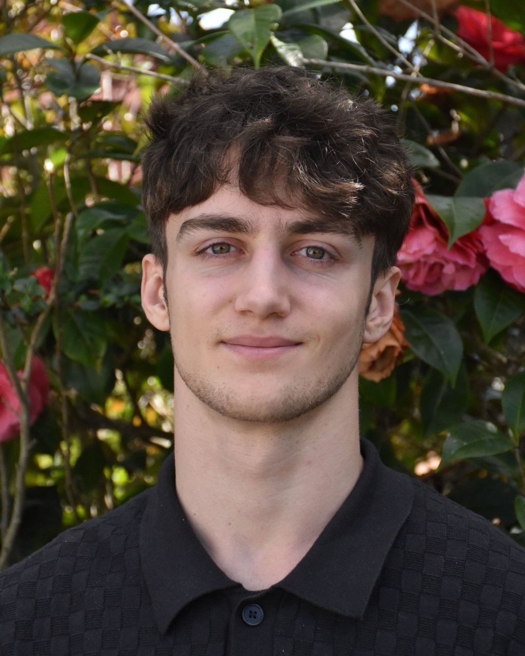 A young man with brown, curly hair and blue eyes standing outdoors in front of flowering bushes with pink flowers.