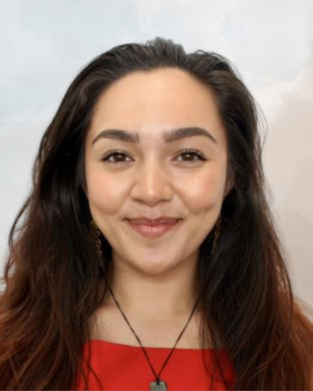 Close-up of a smiling woman with dark brown hair, wearing a red top and a black necklace, standing against a light background.