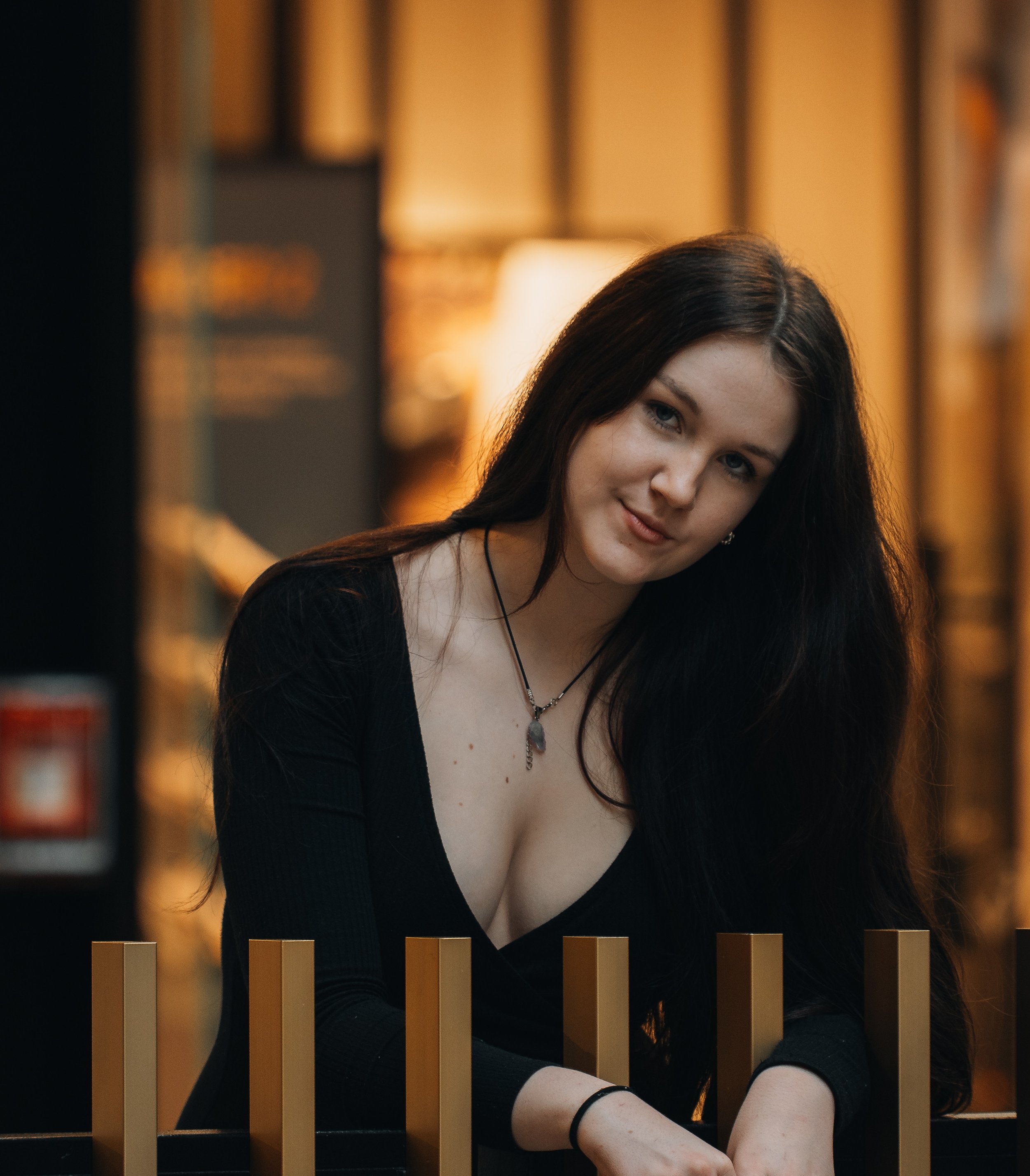A young woman with long dark hair, wearing a black top and a silver necklace, sitting behind a black and gold railing in a warmly lit room.