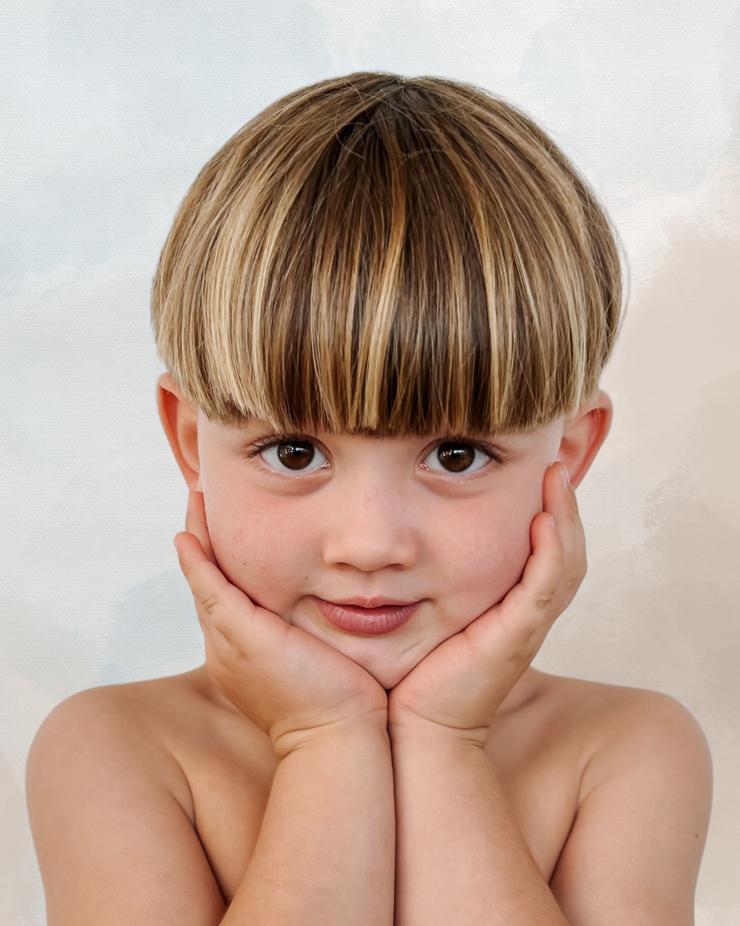 A young boy with light brown hair and brown eyes, looking directly at the camera, resting his face in his hands, smiling softly.