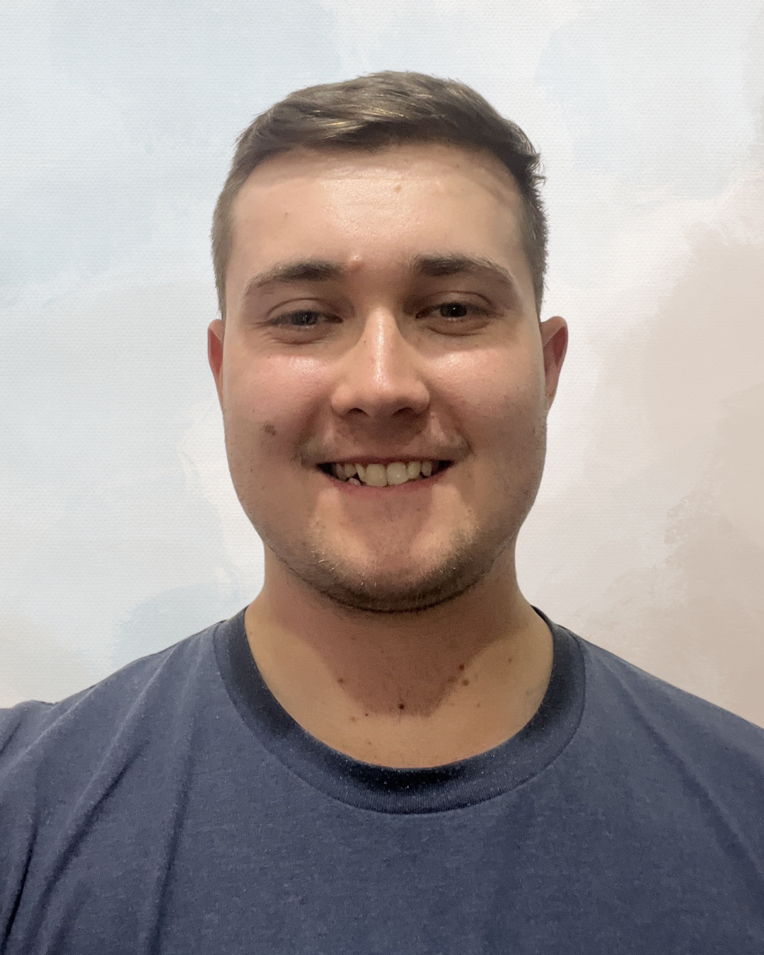A young man with short brown hair smiling at the camera, wearing a dark t-shirt, against a plain light-colored wall.
