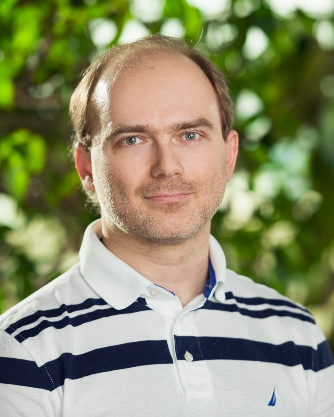 A man with light skin, blue eyes, and brown hair standing outdoors with green foliage in the background, wearing a white polo shirt with navy blue horizontal stripes.