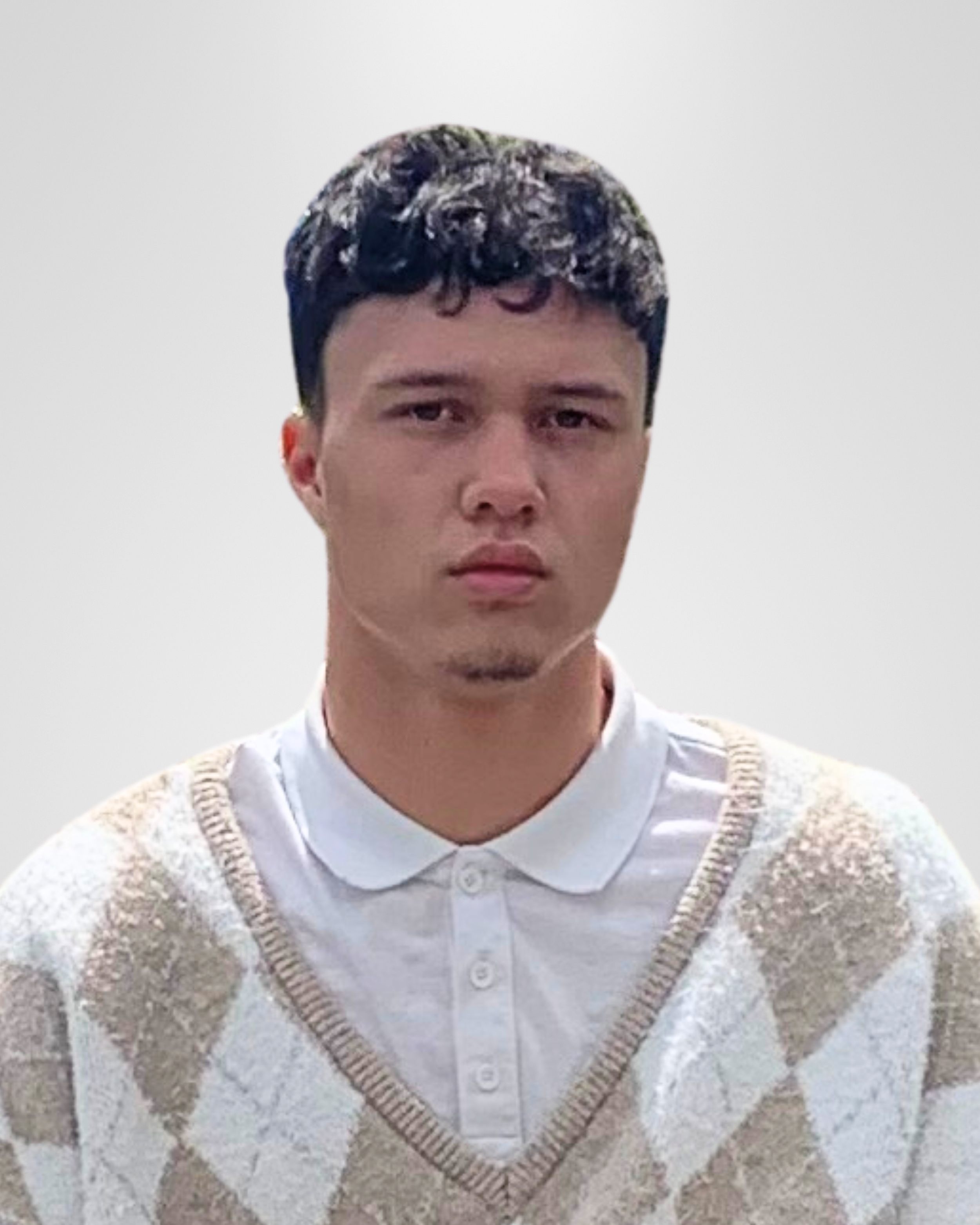 A young man with dark, curly hair wearing a white collared shirt and a beige and white argyle sweater vest, standing against a plain gray background.