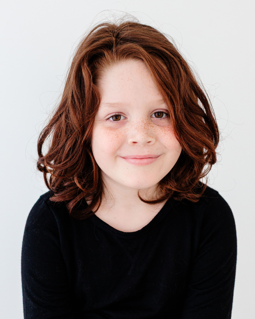 A young girl with red curly hair, freckles, and a gentle smile, wearing a black shirt, against a plain white background.