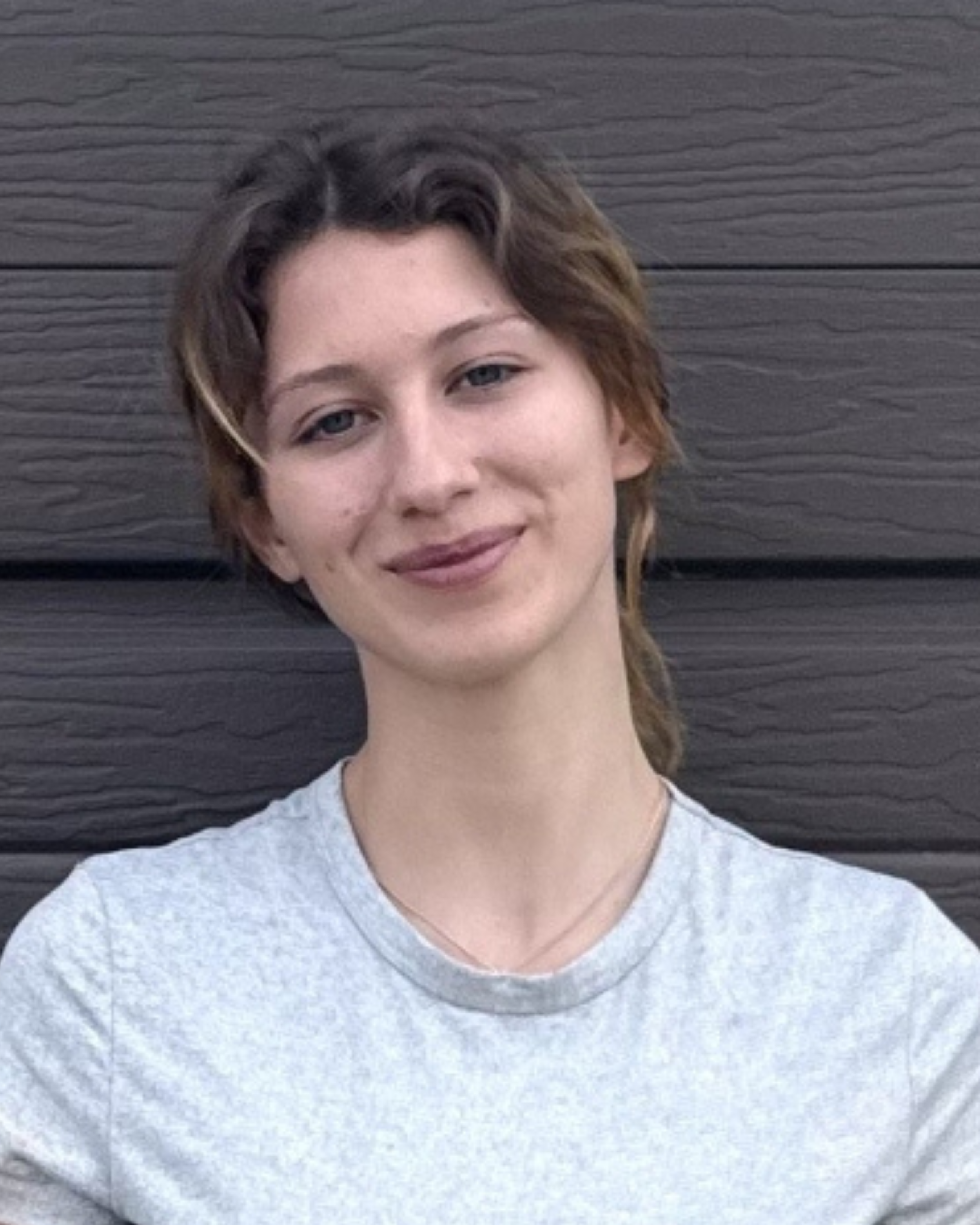 A young woman with short, wavy hair, wearing a gray t-shirt, standing in front of a dark wooden wall, smiling at the camera.
