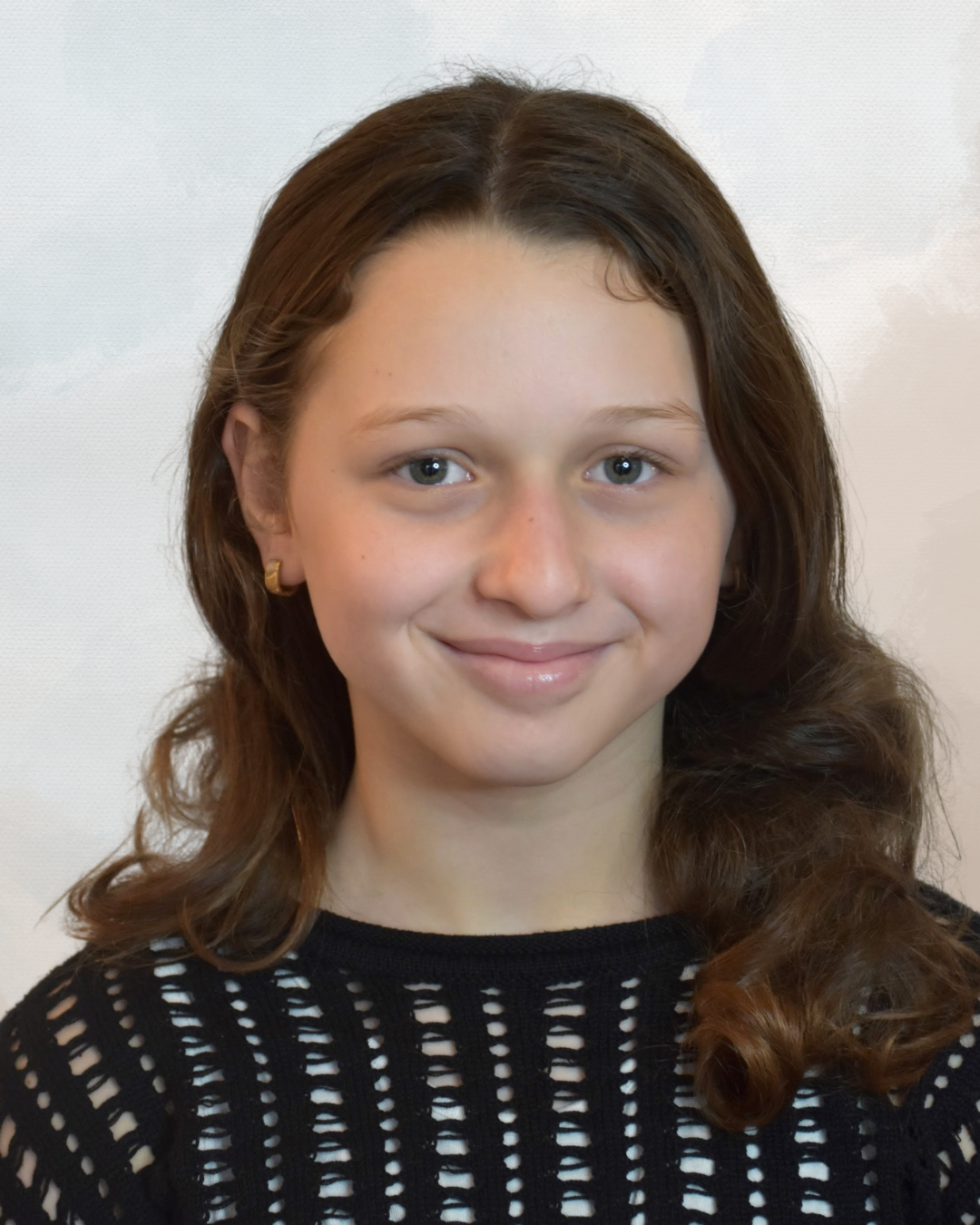 A young woman with wavy brown hair, blue eyes, and light skin, smiling, wearing a black knitted top with cutouts and small gold earrings.