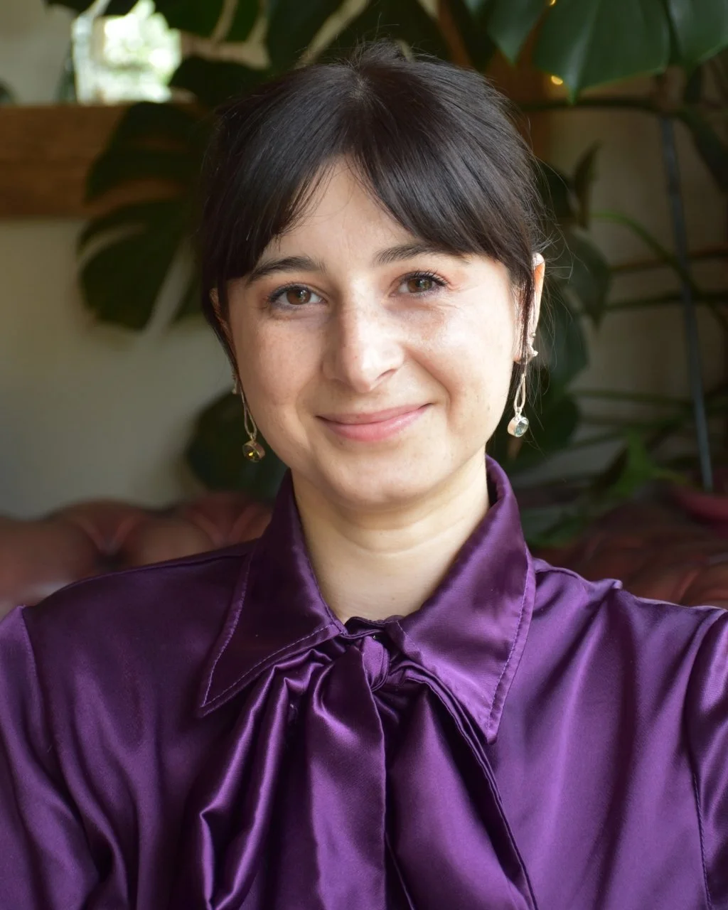 A woman with short dark hair and light skin, smiling, wearing a purple satin blouse with a bow tie, earrings, and sitting indoors with large green plants in the background.
