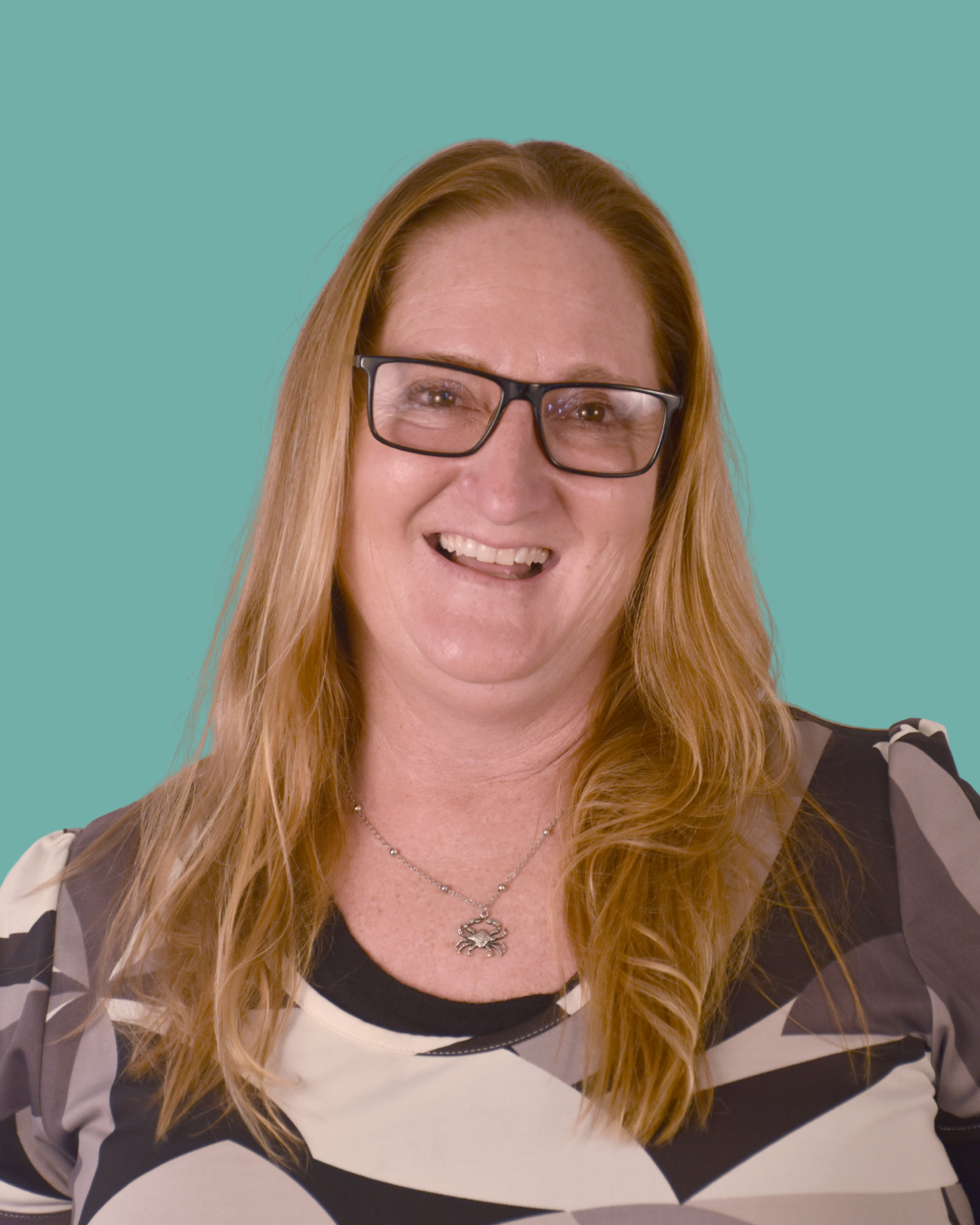 A woman with long red hair, wearing glasses and a black, white, and gray geometric patterned shirt, smiling at the camera with a turquoise background.