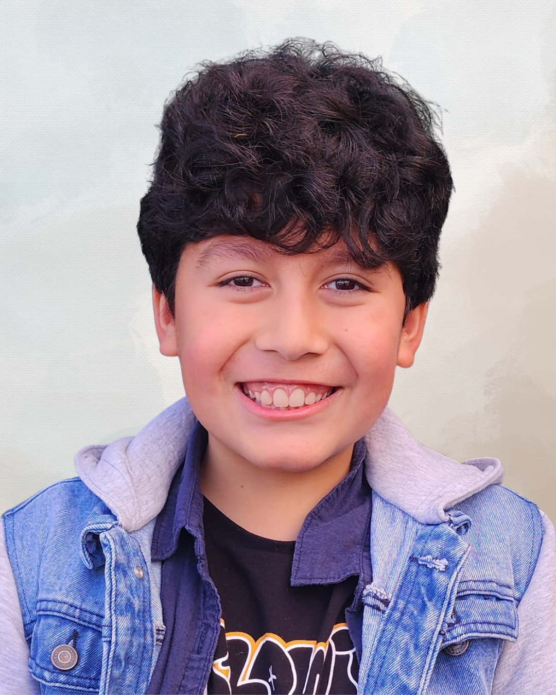 A young boy with curly dark hair smiling, wearing a denim jacket over a black t-shirt, against a light background.
