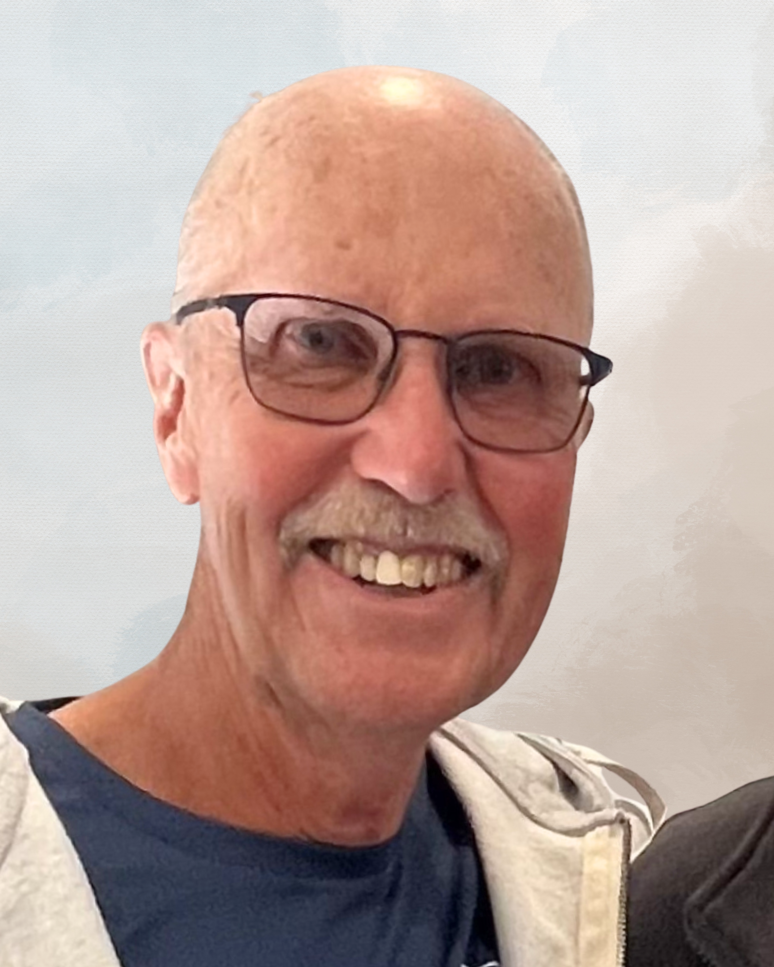Close-up of a smiling man with glasses, bald head, and a mustache, wearing a dark T-shirt and a light-colored hoodie.