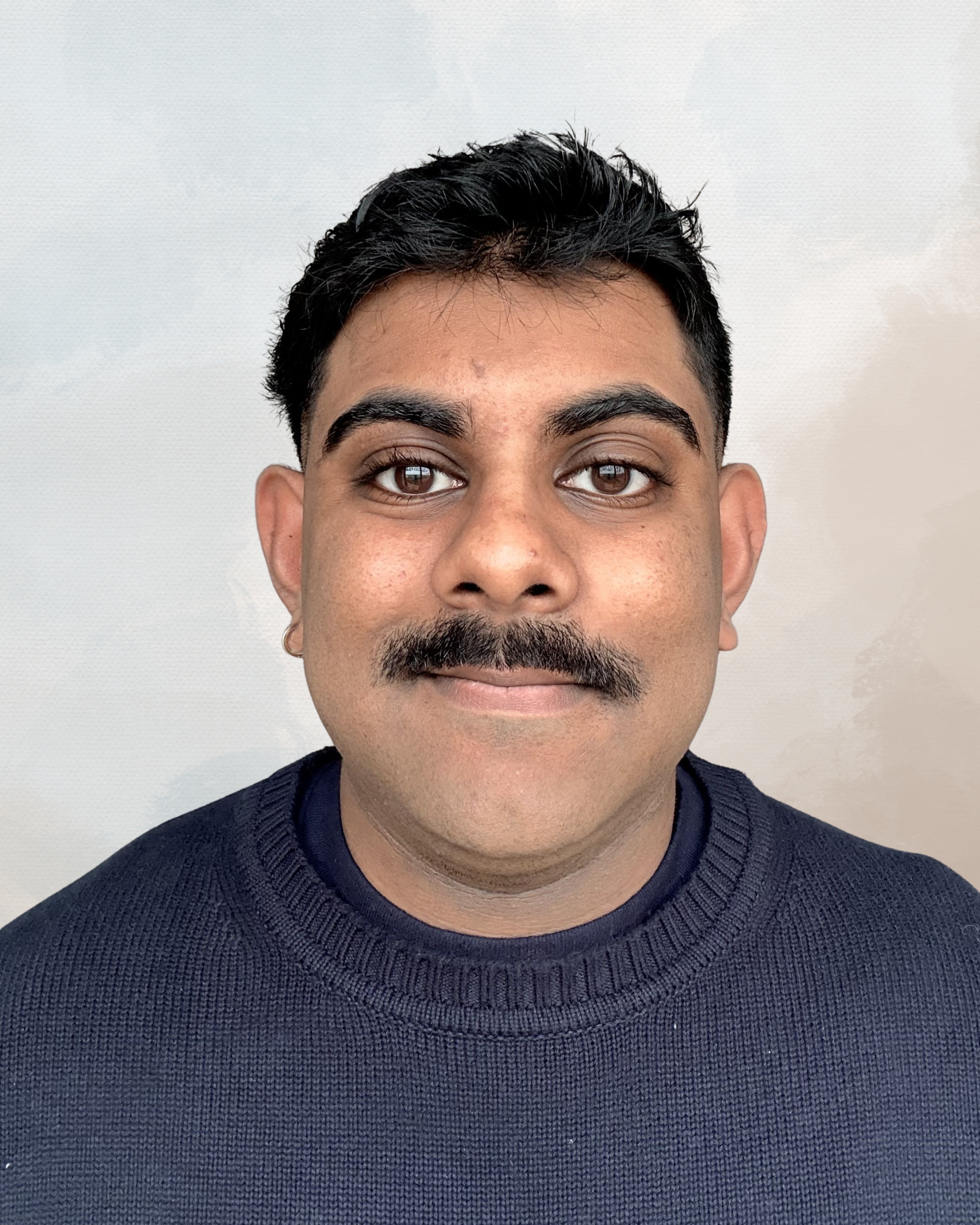 A man with dark hair, brown eyes, and a mustache, wearing a dark blue sweater, standing against a plain white background.