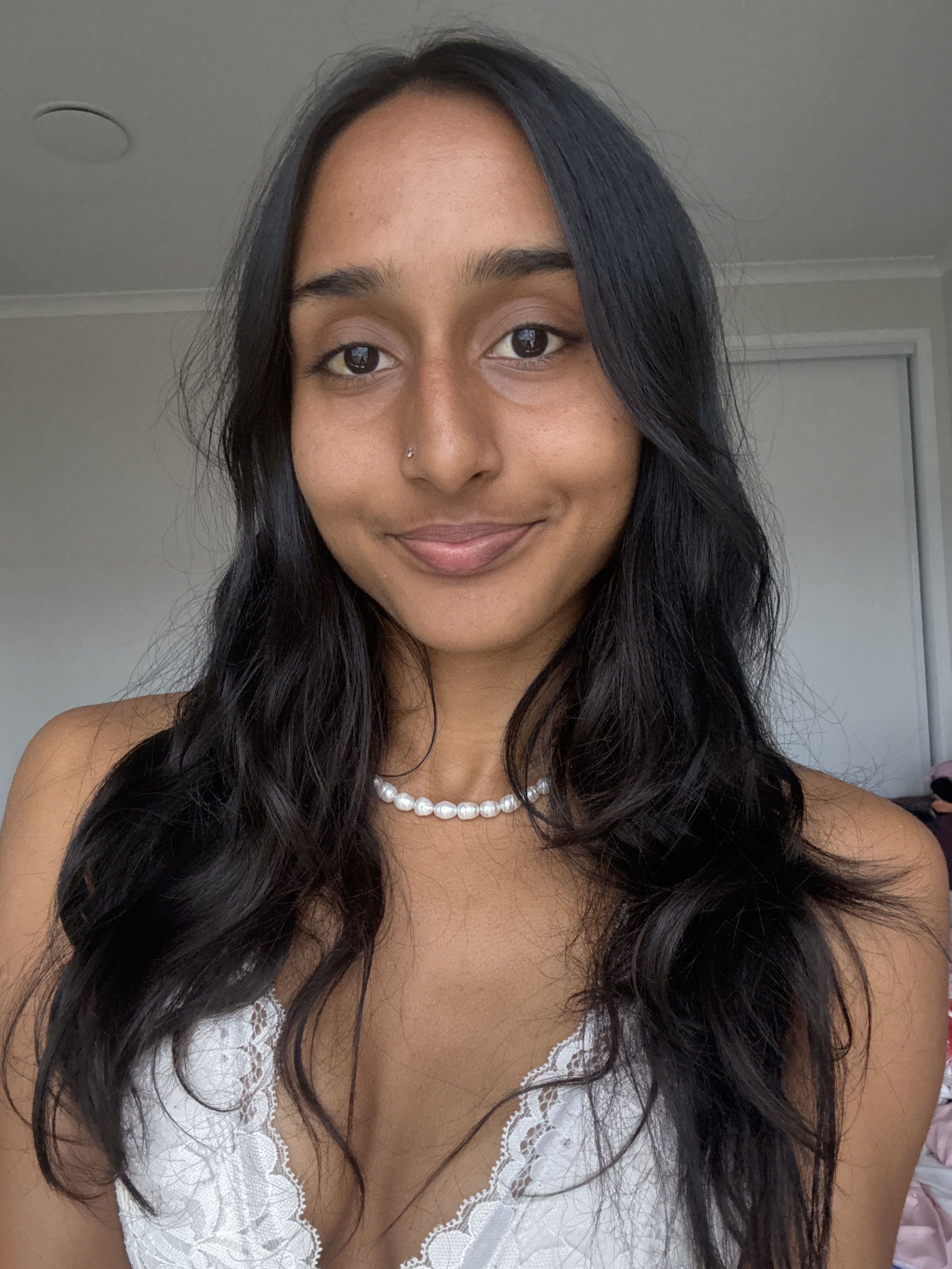 A young woman with long, wavy black hair, wearing a pearl necklace and a white lace top, smiling in a room with neutral-colored walls.