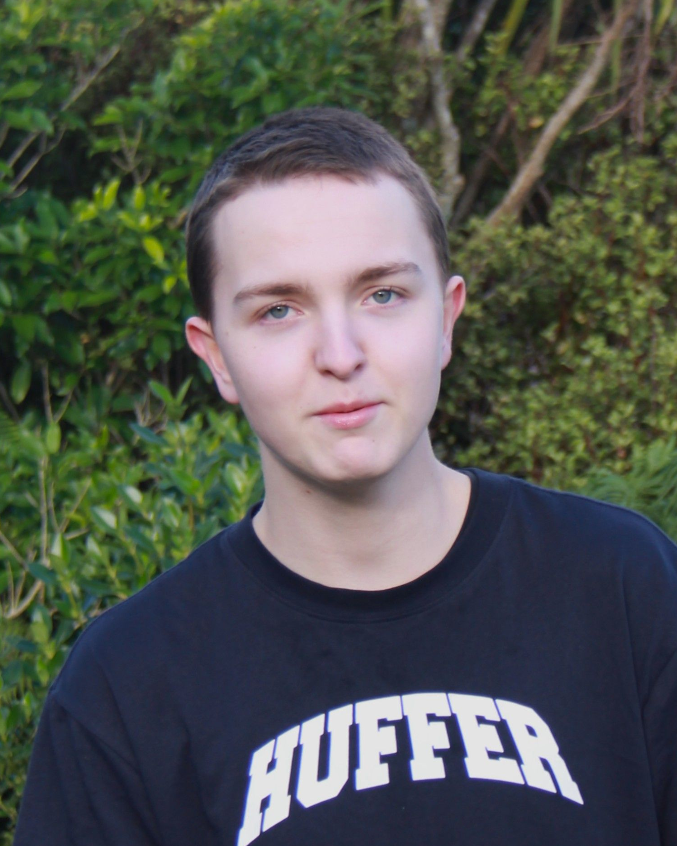 Young man with short brown hair and blue eyes standing outdoors in front of green foliage, wearing a black shirt with the word 'HUFFER' printed on it.