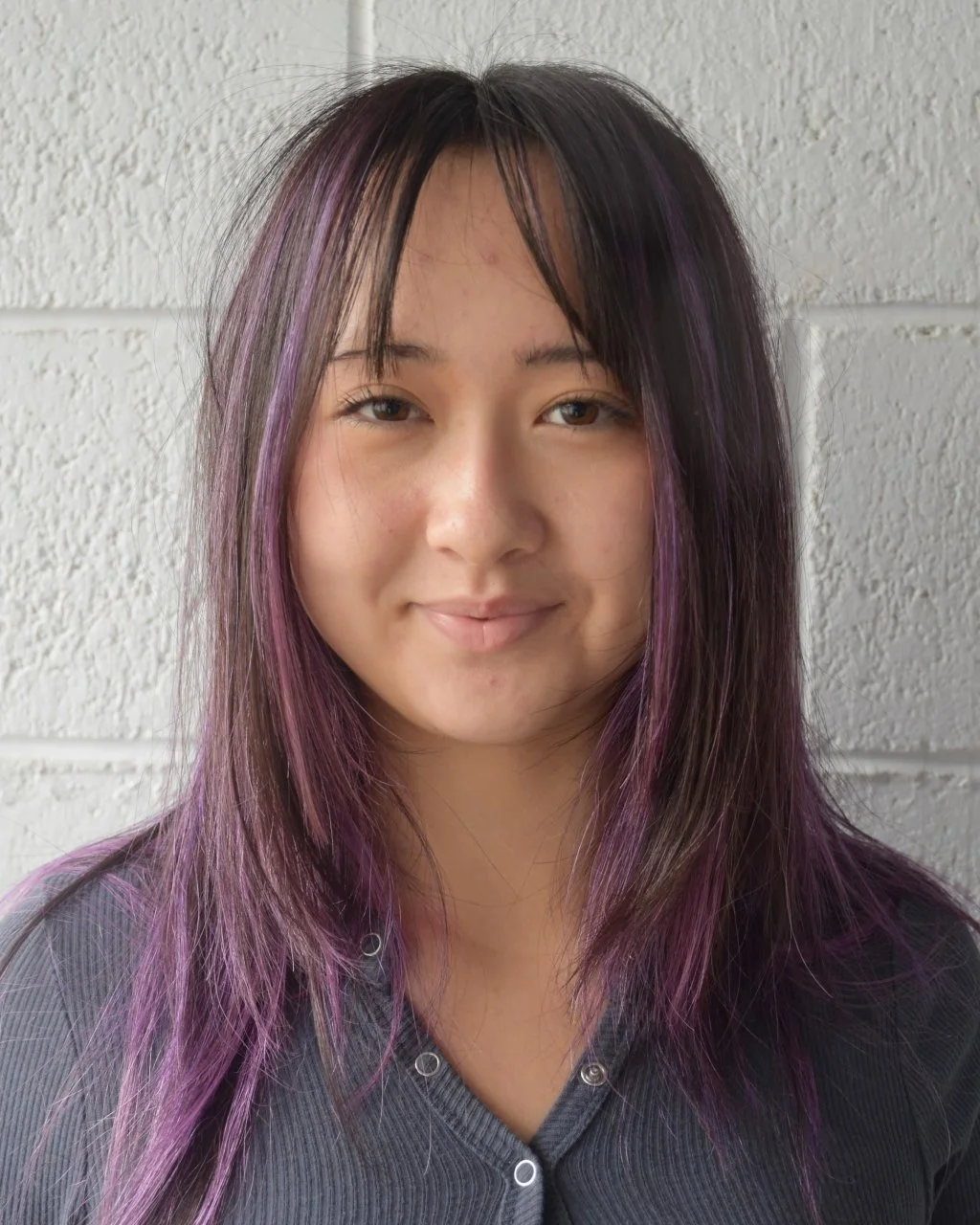 Portrait of a young woman with shoulder-length dark hair with purple highlights, standing against a light gray textured wall, wearing a dark gray top.