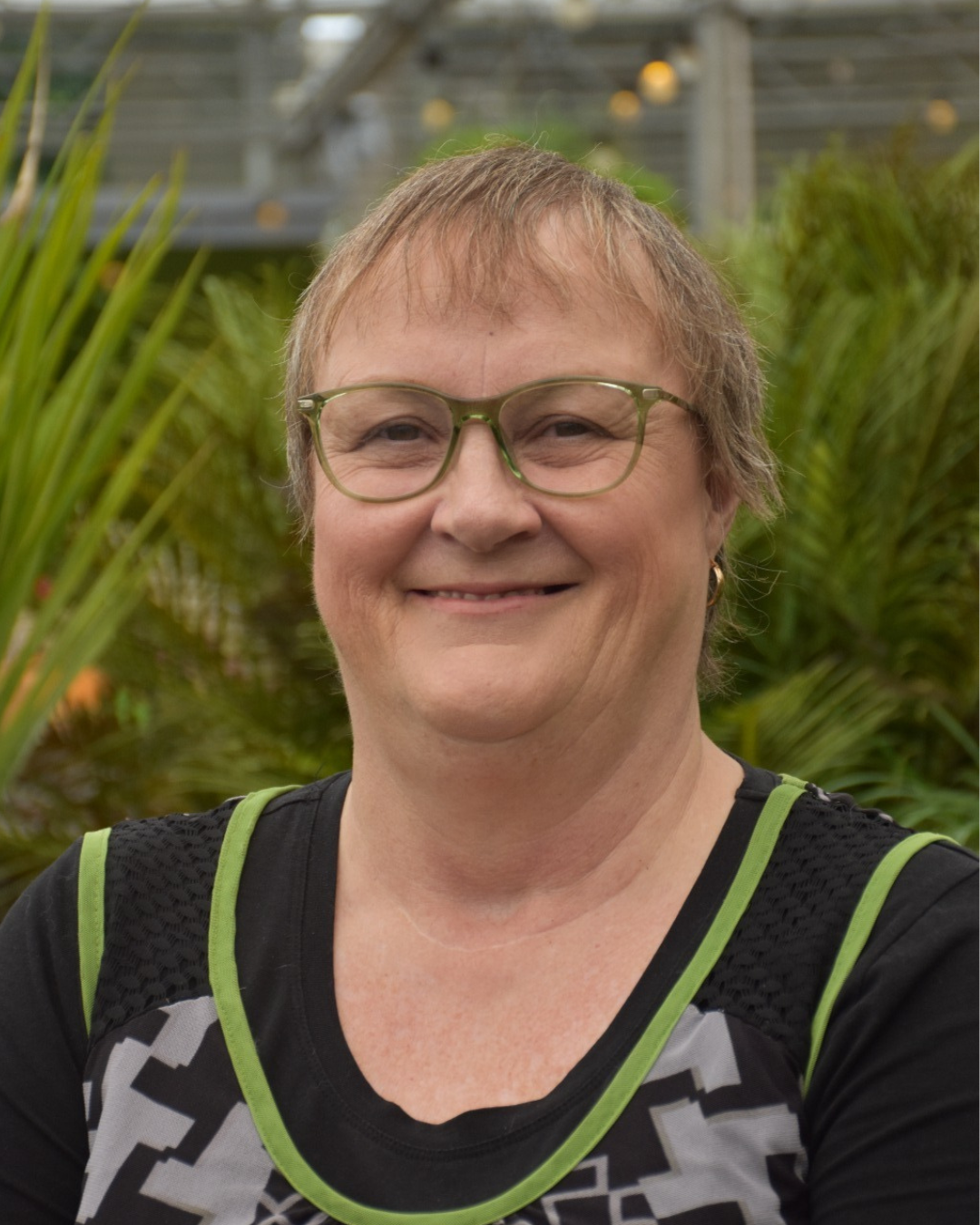 A smiling woman with short blond hair, glasses, wearing a black and gray athletic shirt with green accents, standing outdoors with green plants in the background.