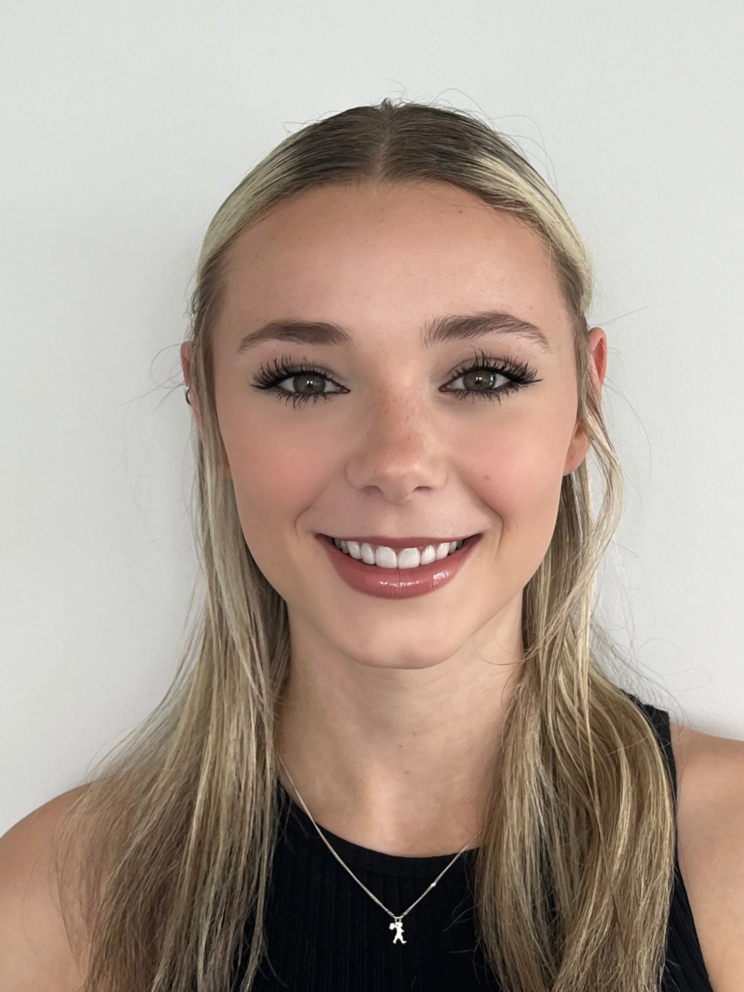 A young woman with long blonde hair, wearing a black sleeveless top and a silver necklace with a teddy bear pendant, smiling in front of a plain white wall.