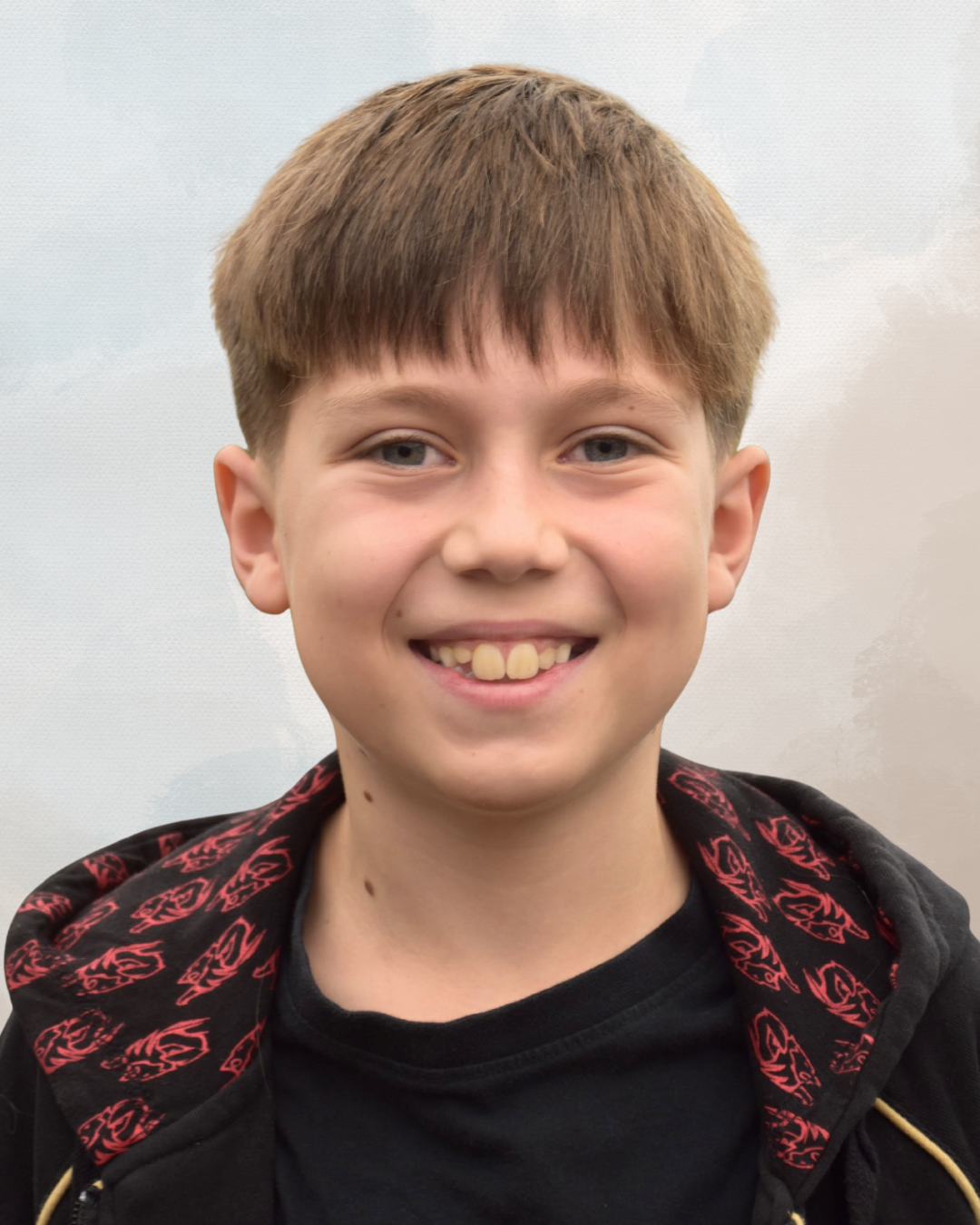Young boy with light brown hair, smiling, wearing a black shirt and a black jacket with red designs, standing outdoors against a cloudy sky.