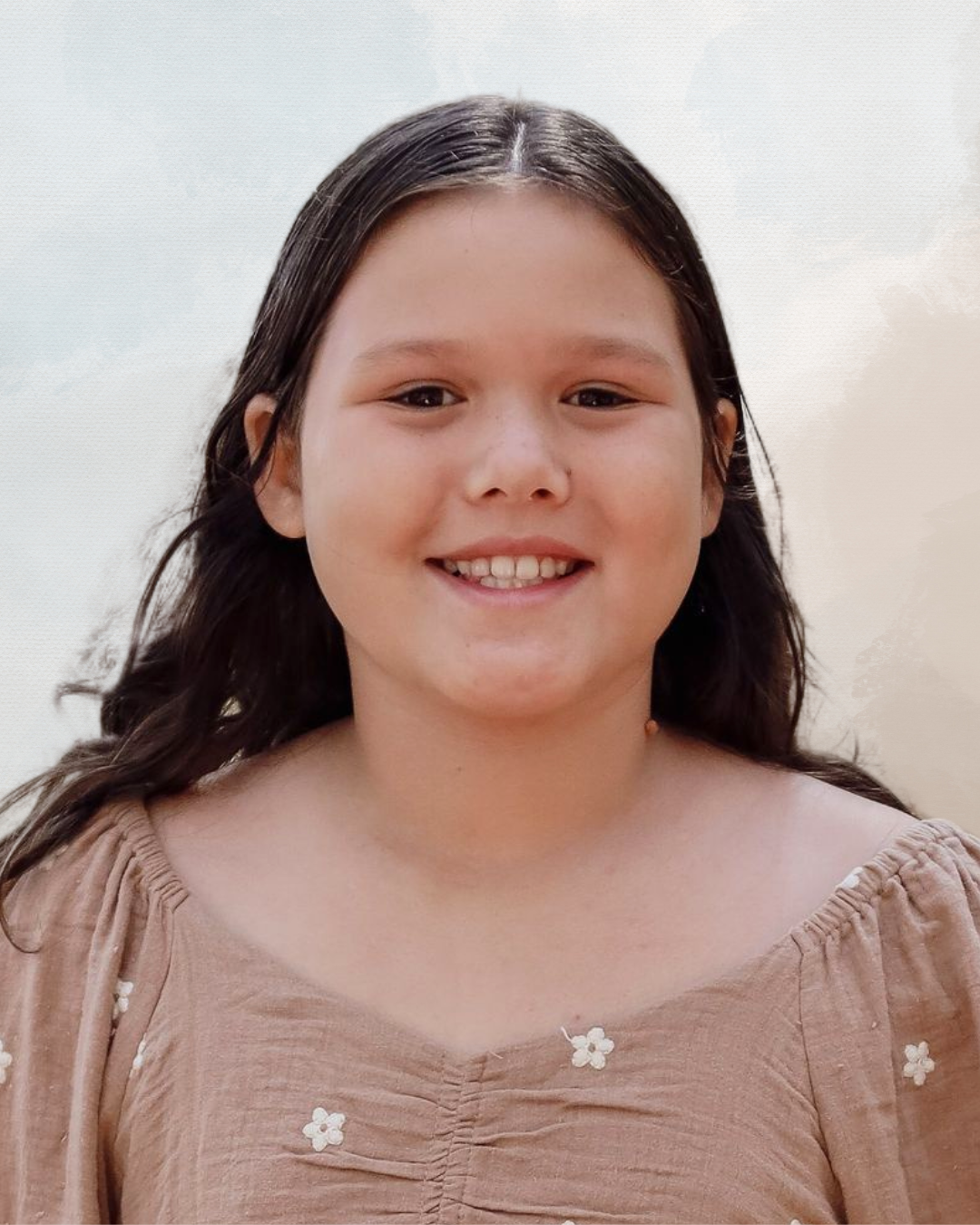 Young girl with brown hair smiling, wearing a beige dress with white flower embroidery, outdoors against a light sky background.