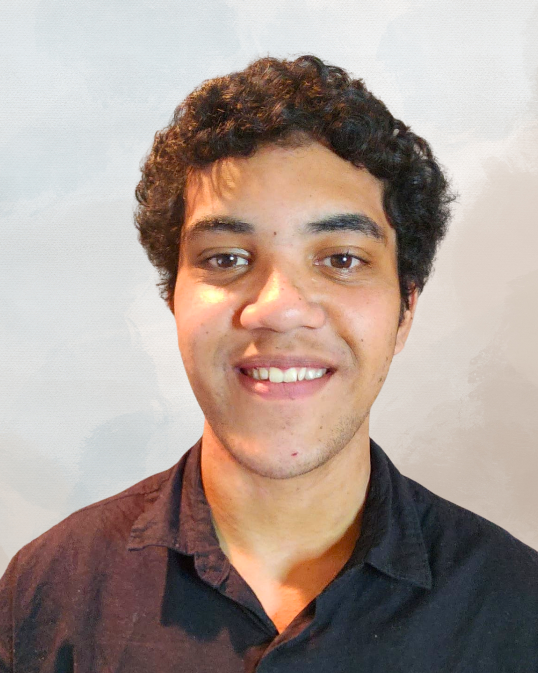 Close-up portrait of a smiling young man with dark curly hair wearing a black shirt, standing against a light neutral background.