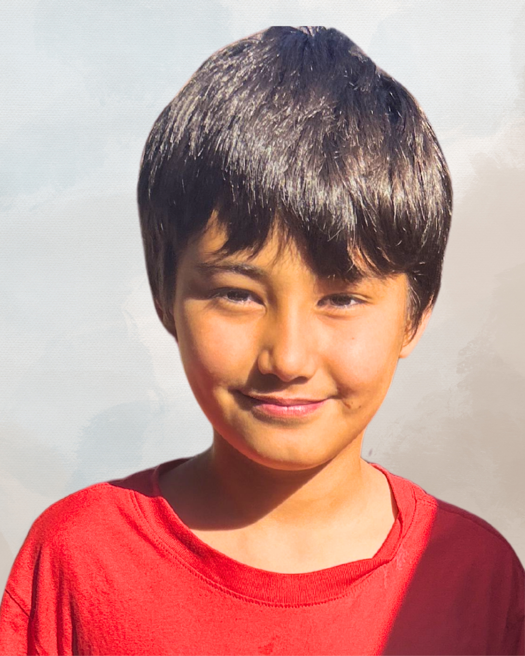 Close-up of a young boy with straight dark hair, wearing a red shirt, smiling softly, against a neutral background.