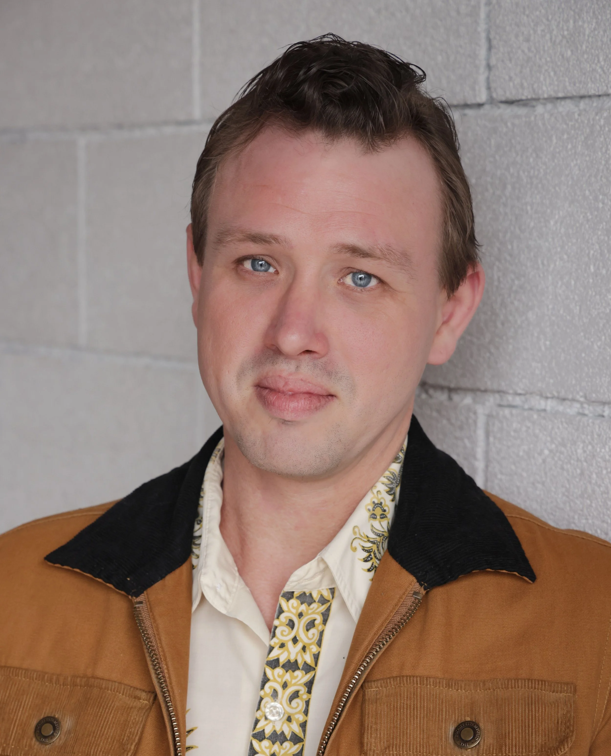 Close-up of a man with blue eyes and short brown hair standing against a gray brick wall, wearing a beige shirt with a patterned collar and a brown jacket with black collar details.