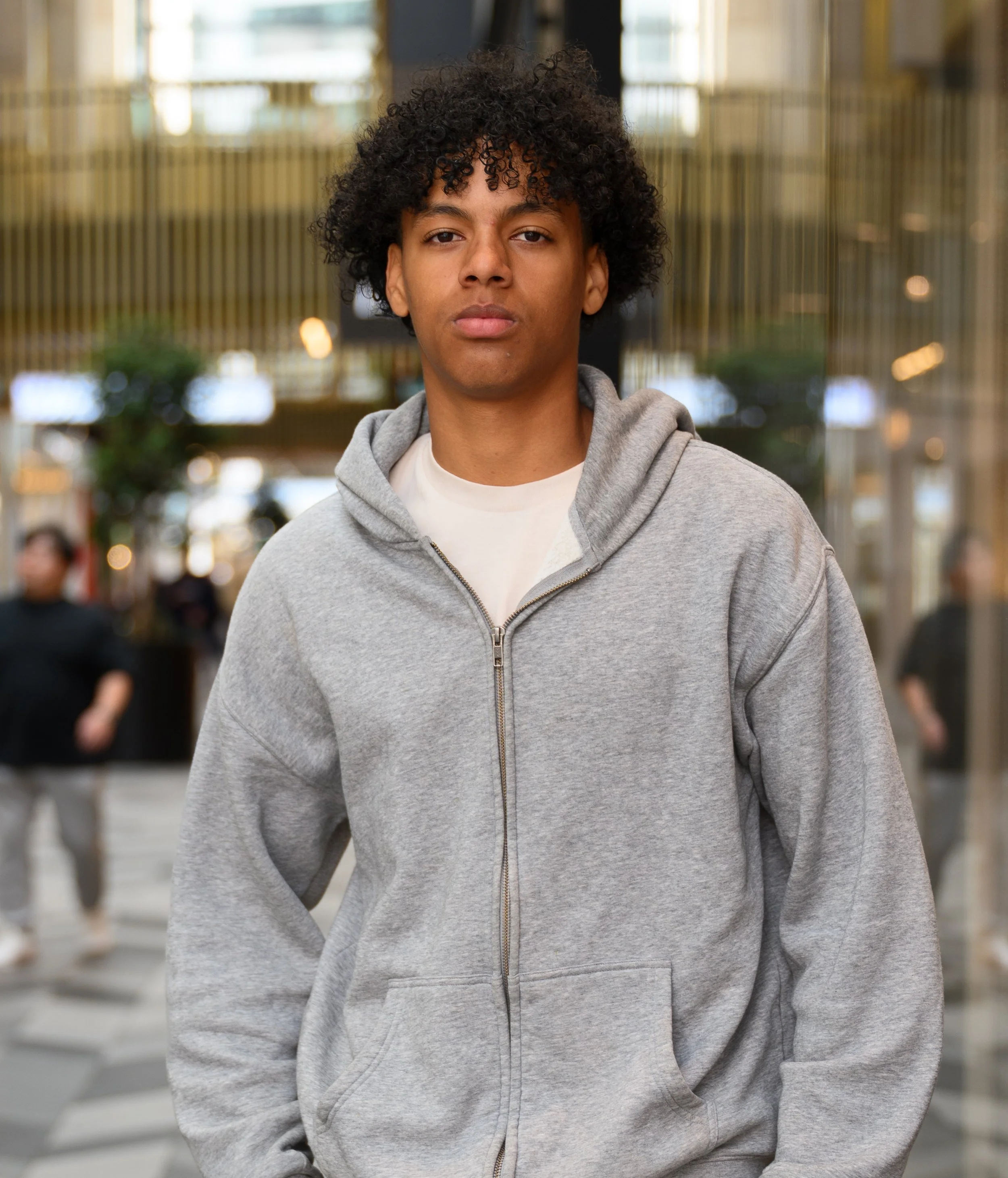Young man with curly hair wearing a gray hoodie in an urban setting, with blurred individuals and buildings in the background.