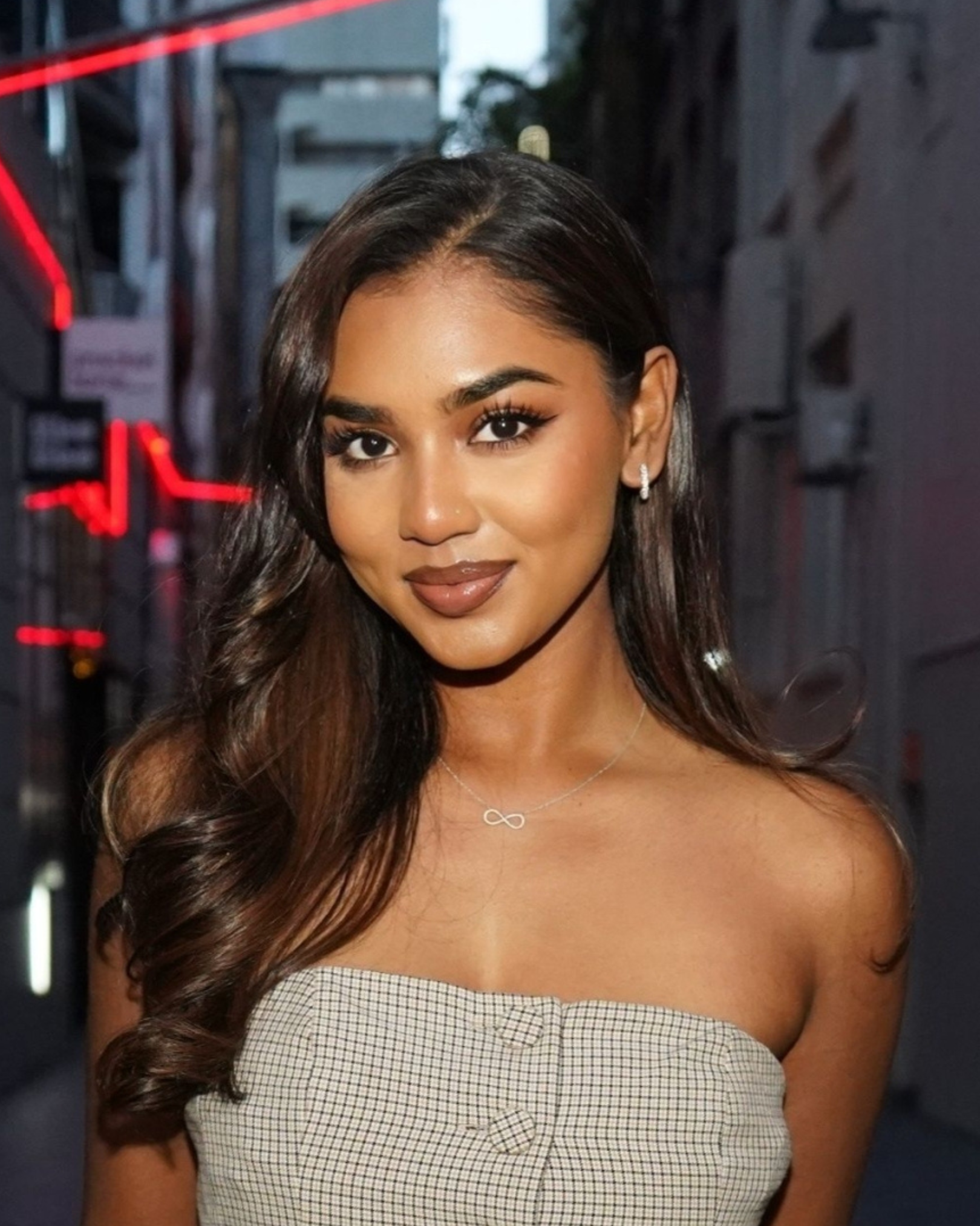 A young woman with long, wavy dark hair smiling outdoors in the evening, wearing a strapless checkered top, hoop earrings, and a necklace with an infinity symbol.