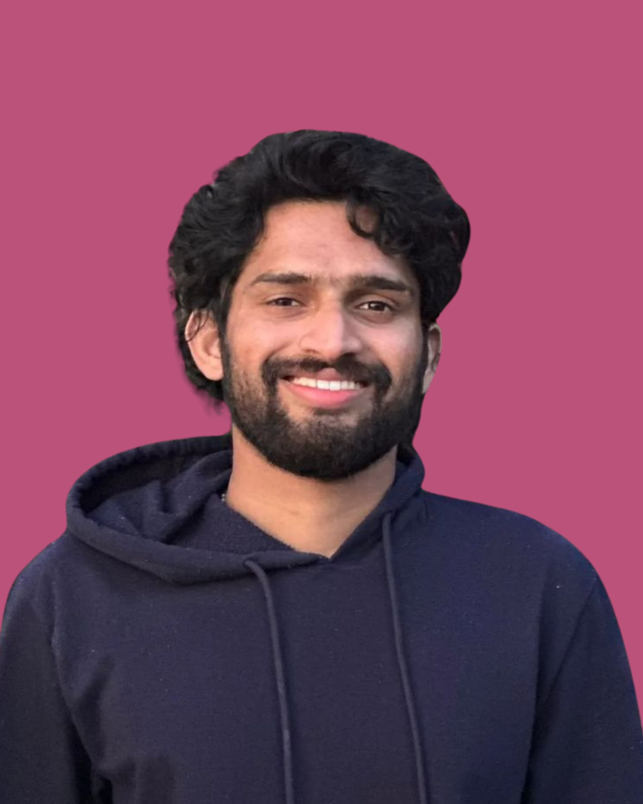 Young man with dark, curly hair and a beard, smiling while wearing a dark hoodie against a pink background.