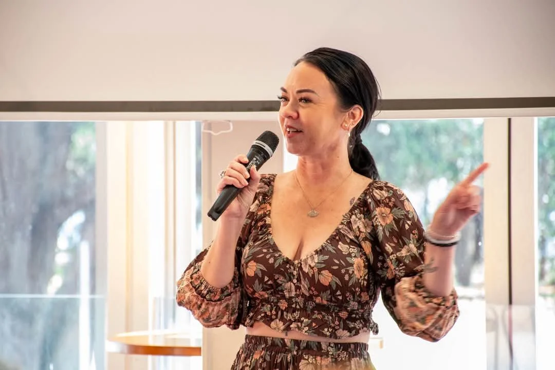 Woman speaking into a microphone during a presentation, standing in front of a window with trees outside.
