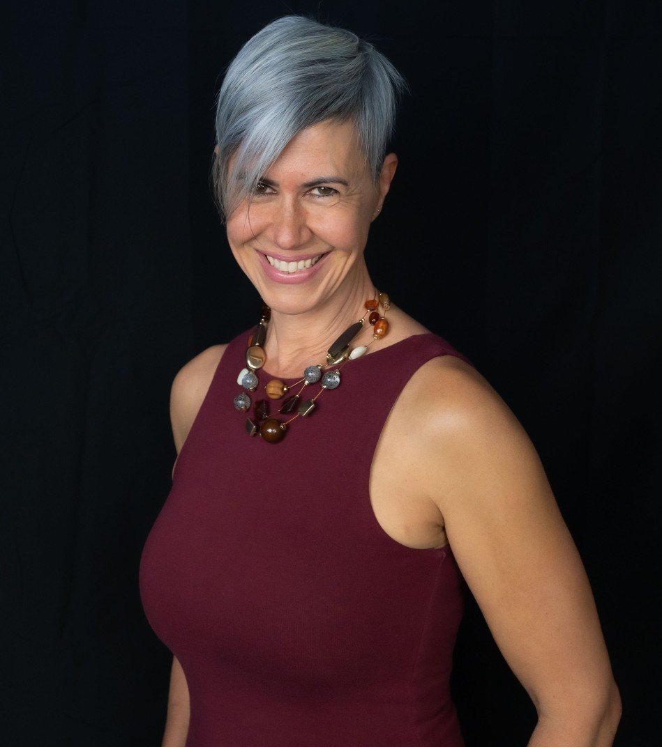 A woman with short gray hair wearing a sleeveless maroon top and a multicolored beaded necklace, smiling against a black background.