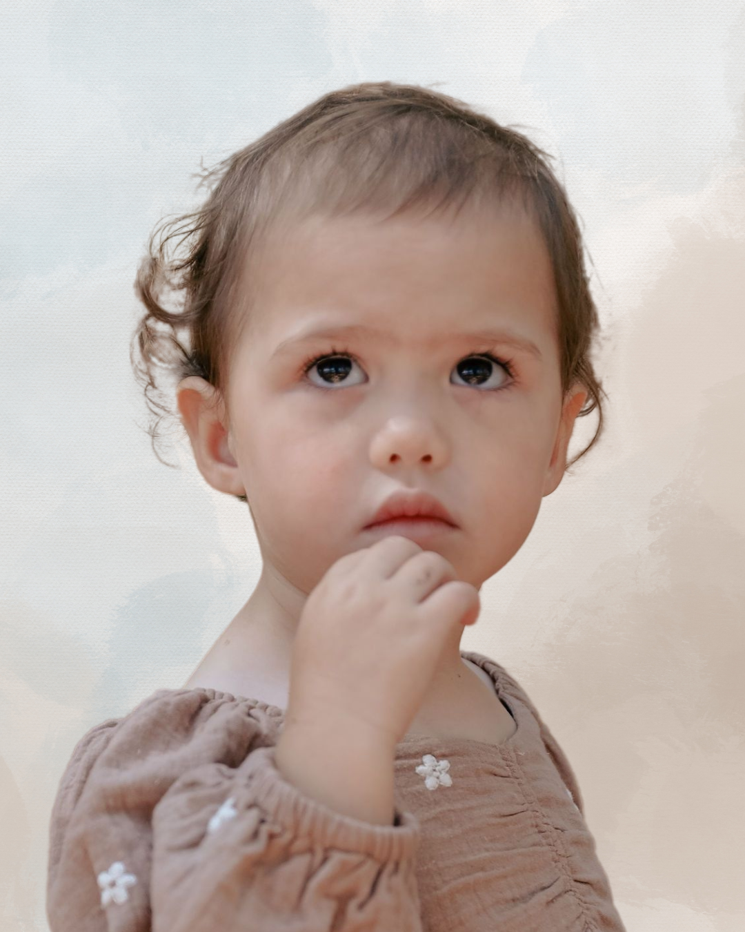 A young child with curly brown hair and dark eyes, wearing a brown top with small white flower embroidery, looks upward with a thoughtful expression, hand near their mouth.
