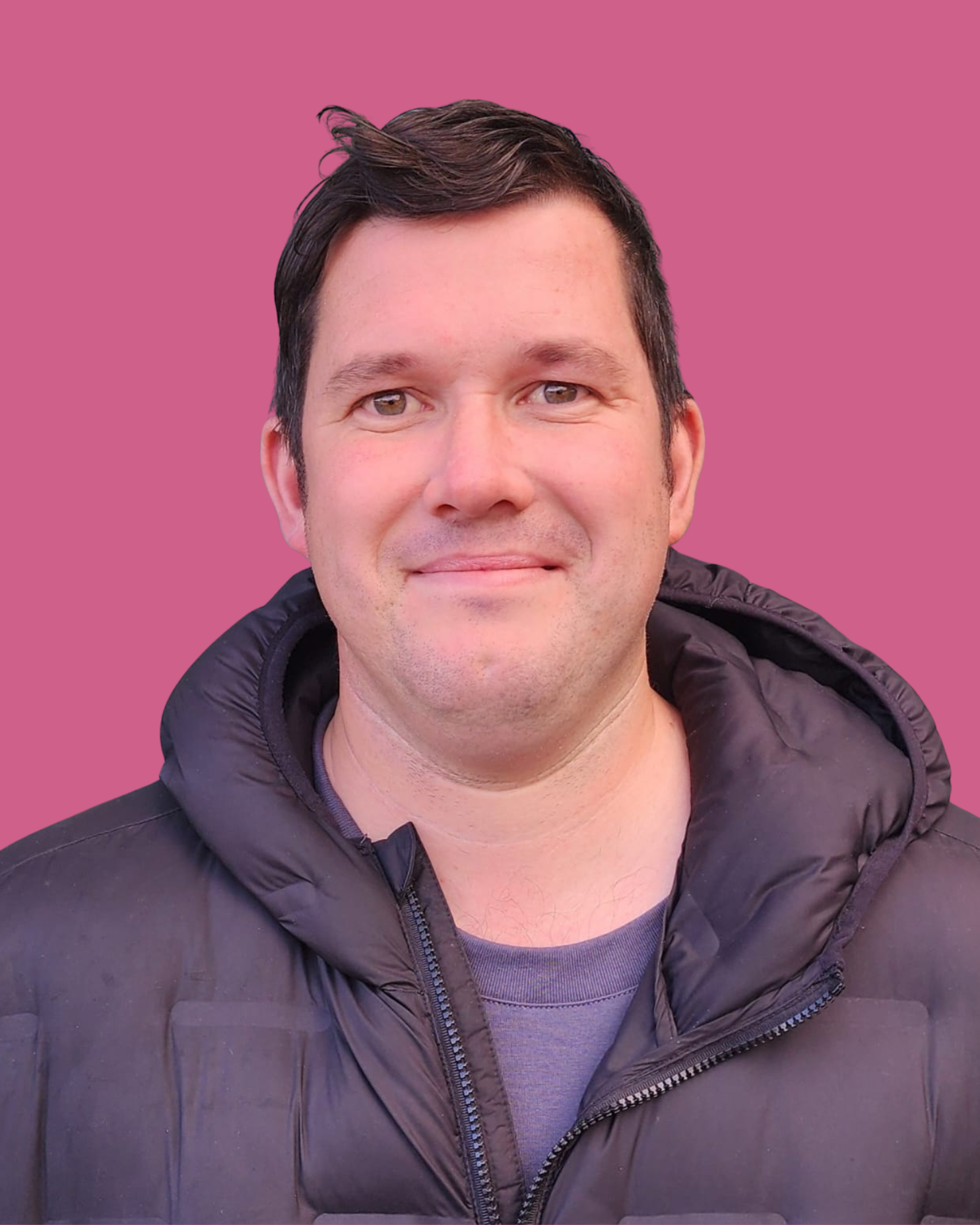 Portrait of a man with short dark hair, wearing a black puffy jacket and a gray shirt, standing against a pink background.