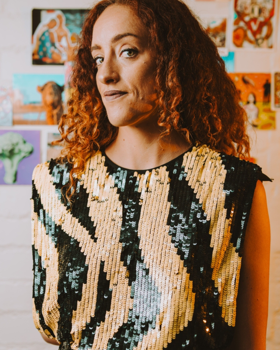 A woman with curly red hair wearing a black and gold sequined top standing in front of colorful artwork on a wall.