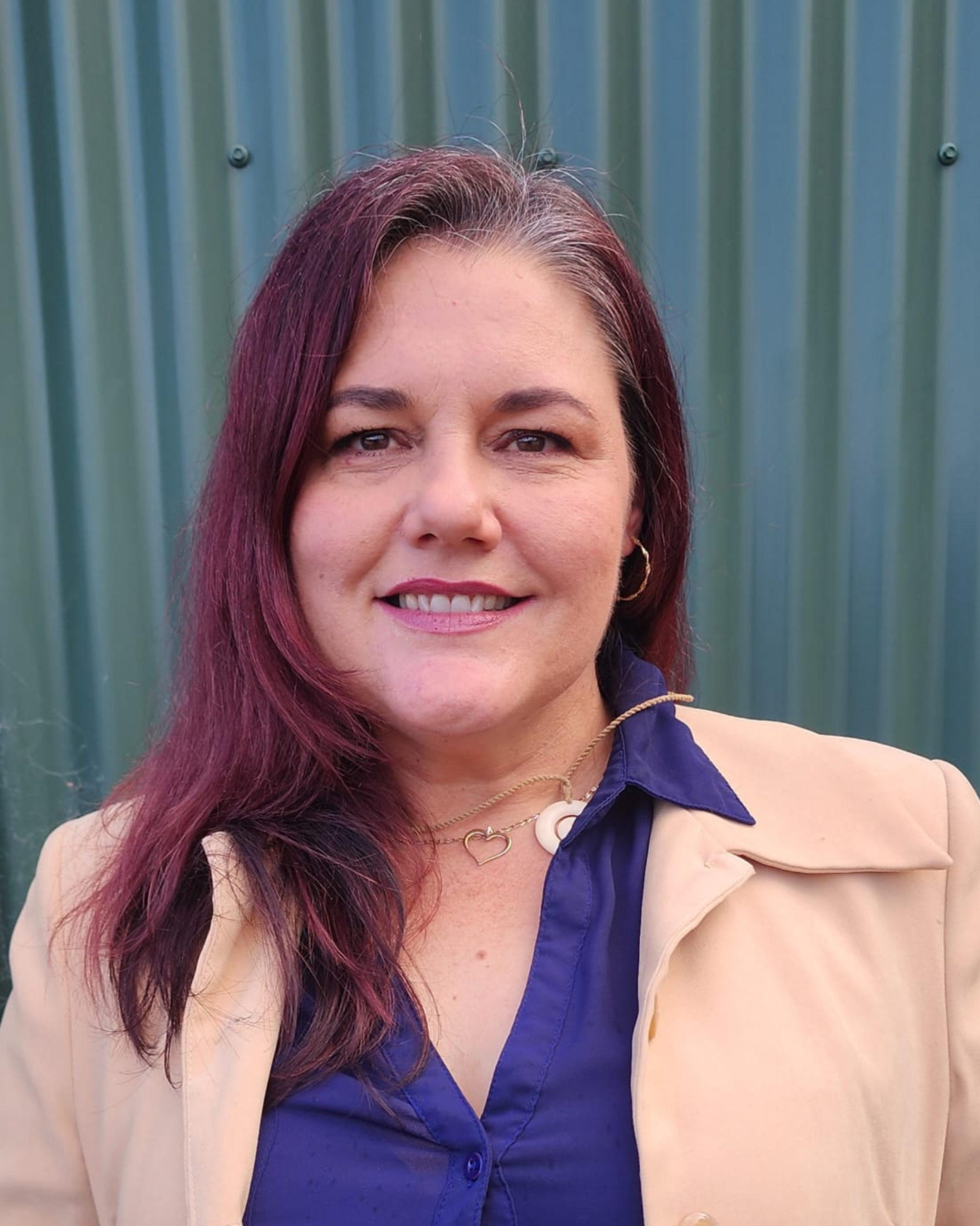 A woman with shoulder-length dark red hair, wearing a beige blazer over a navy blue shirt, standing in front of a green corrugated metal wall.
