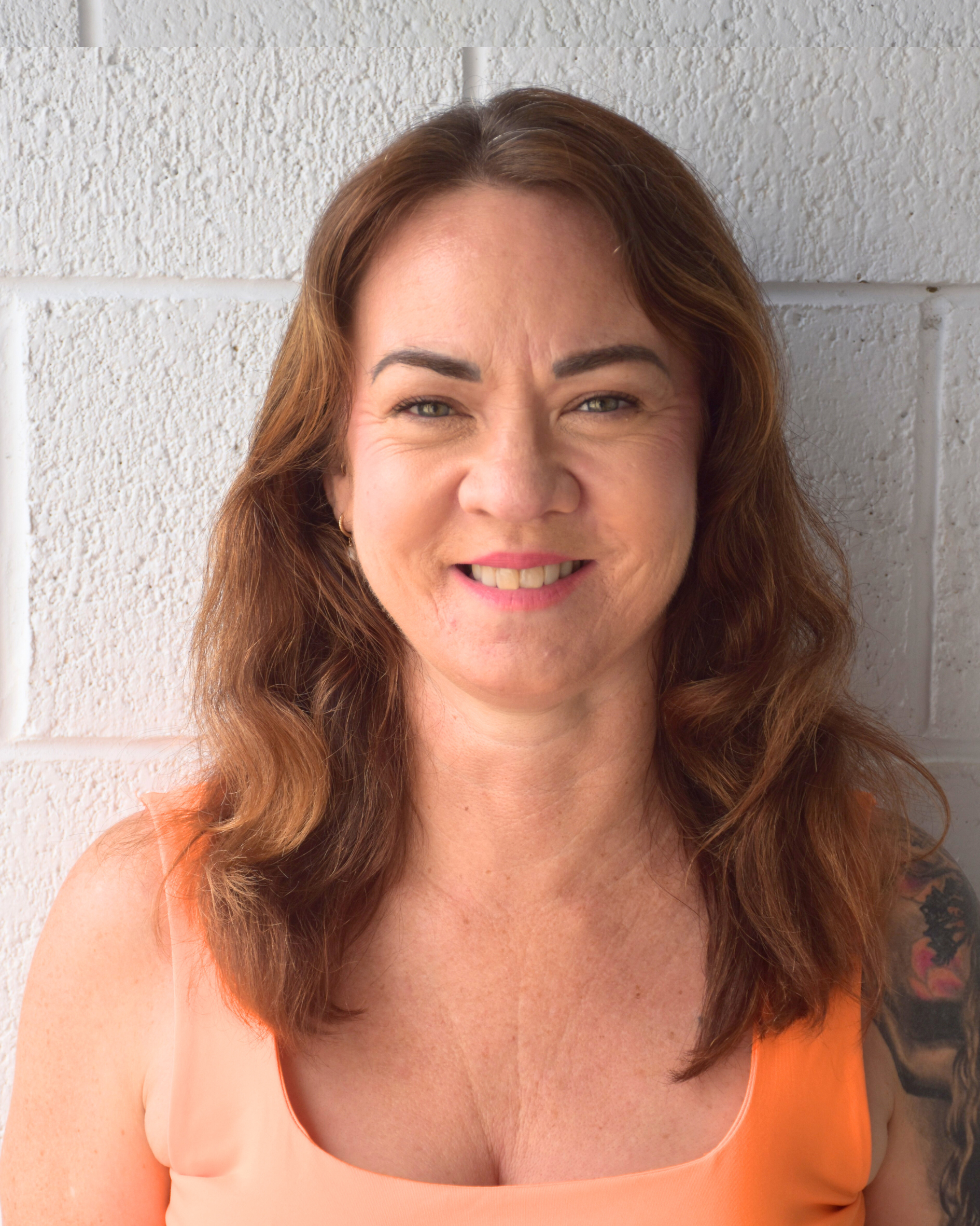 Close-up of a woman with wavy brown hair and a tattoo on her right shoulder, smiling against a white brick wall background.