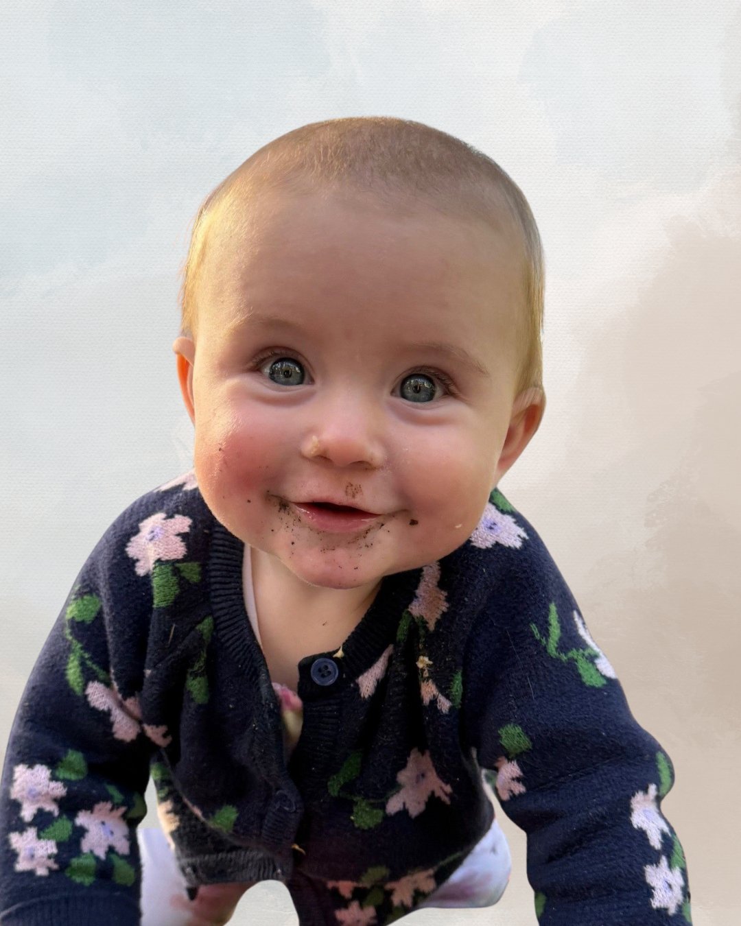 A smiling baby with blue eyes, dirty face, and wearing a navy sweater with pink and green floral pattern.