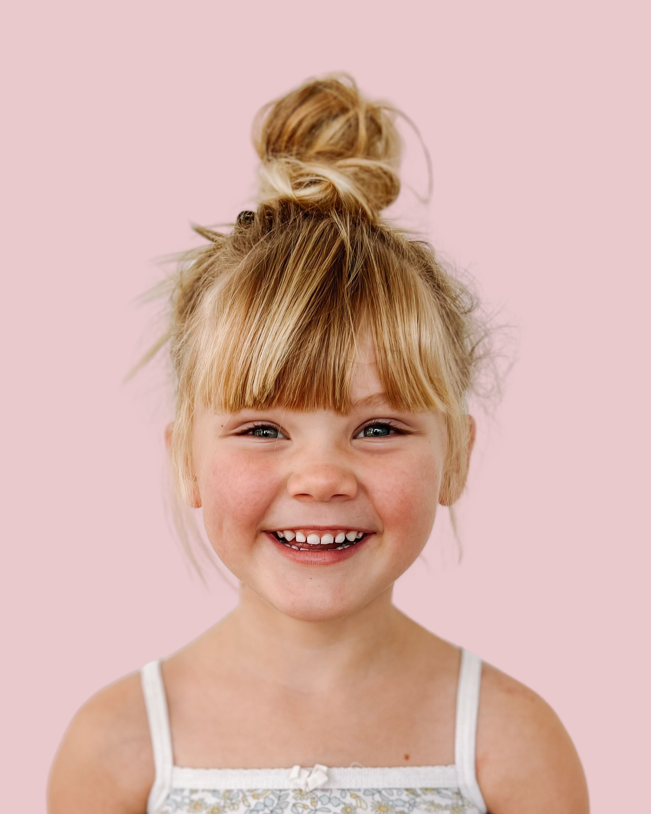 A young girl with red hair styled in a messy top knot and straight bangs, smiling against a pink background.