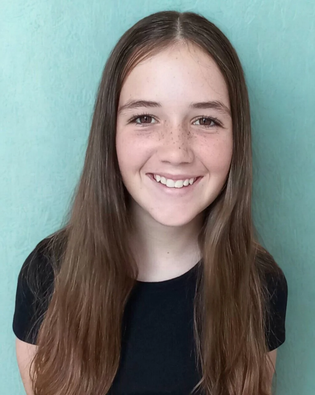 Portrait of a young girl with long brown hair, wearing a white T-shirt with black trim and small earrings.