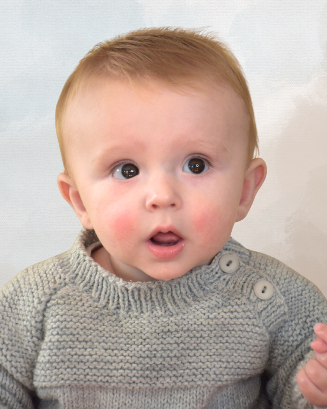 Close-up of a surprised baby with wide eyes, wearing a gray knitted sweater with buttons, against a plain light background.