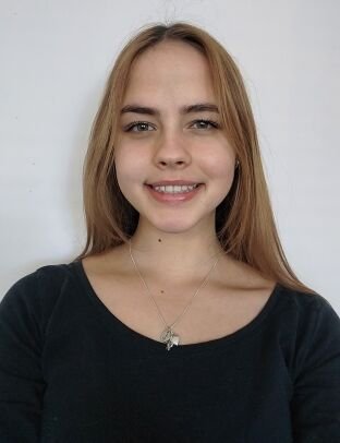 A young woman with long, straight, light brown hair smiling at the camera, wearing a black top and a necklace with a small pendant, against a plain light-colored background.