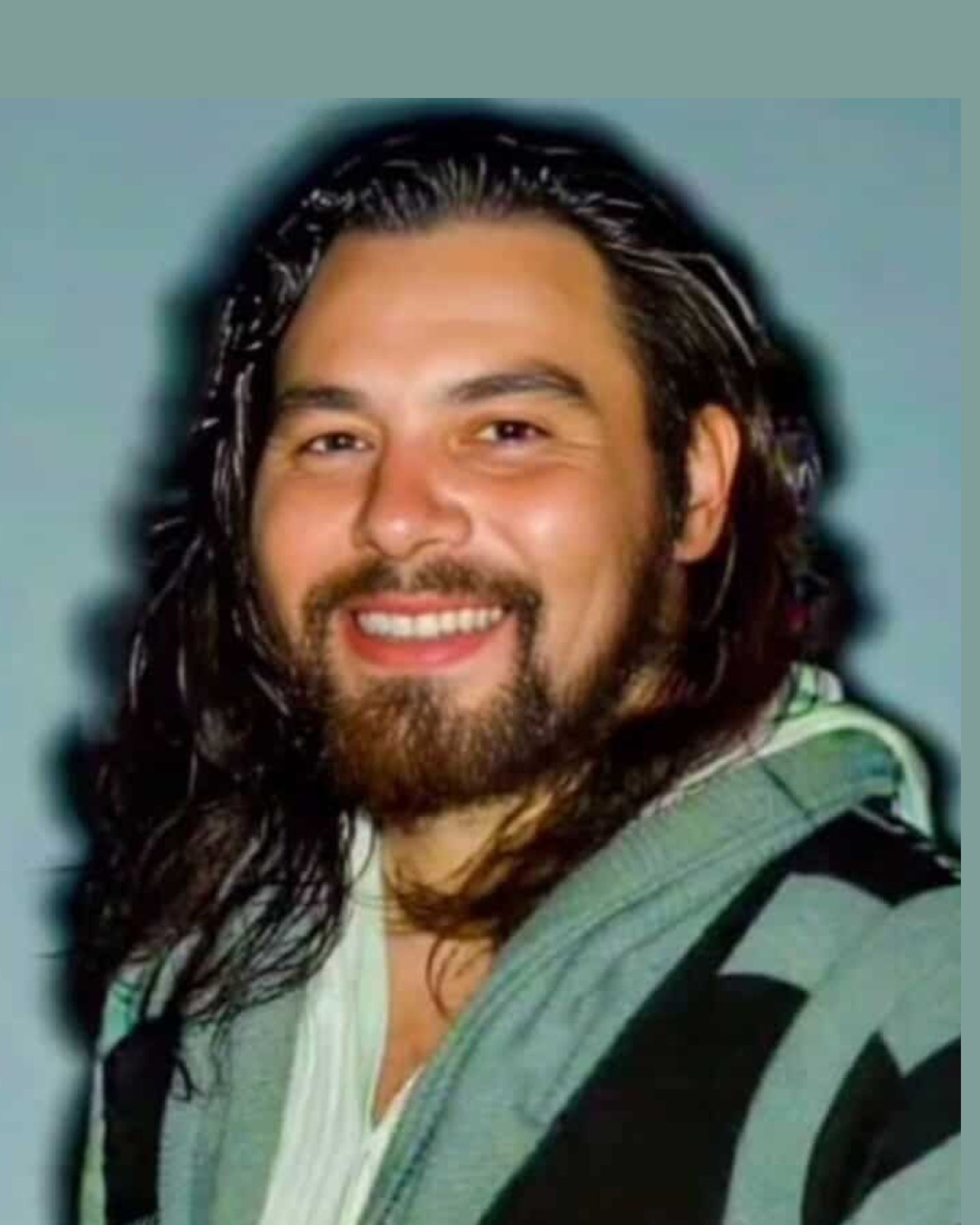A man with long dark hair, a beard, and a mustache smiling at the camera, wearing a patterned shirt and carrying a backpack.
