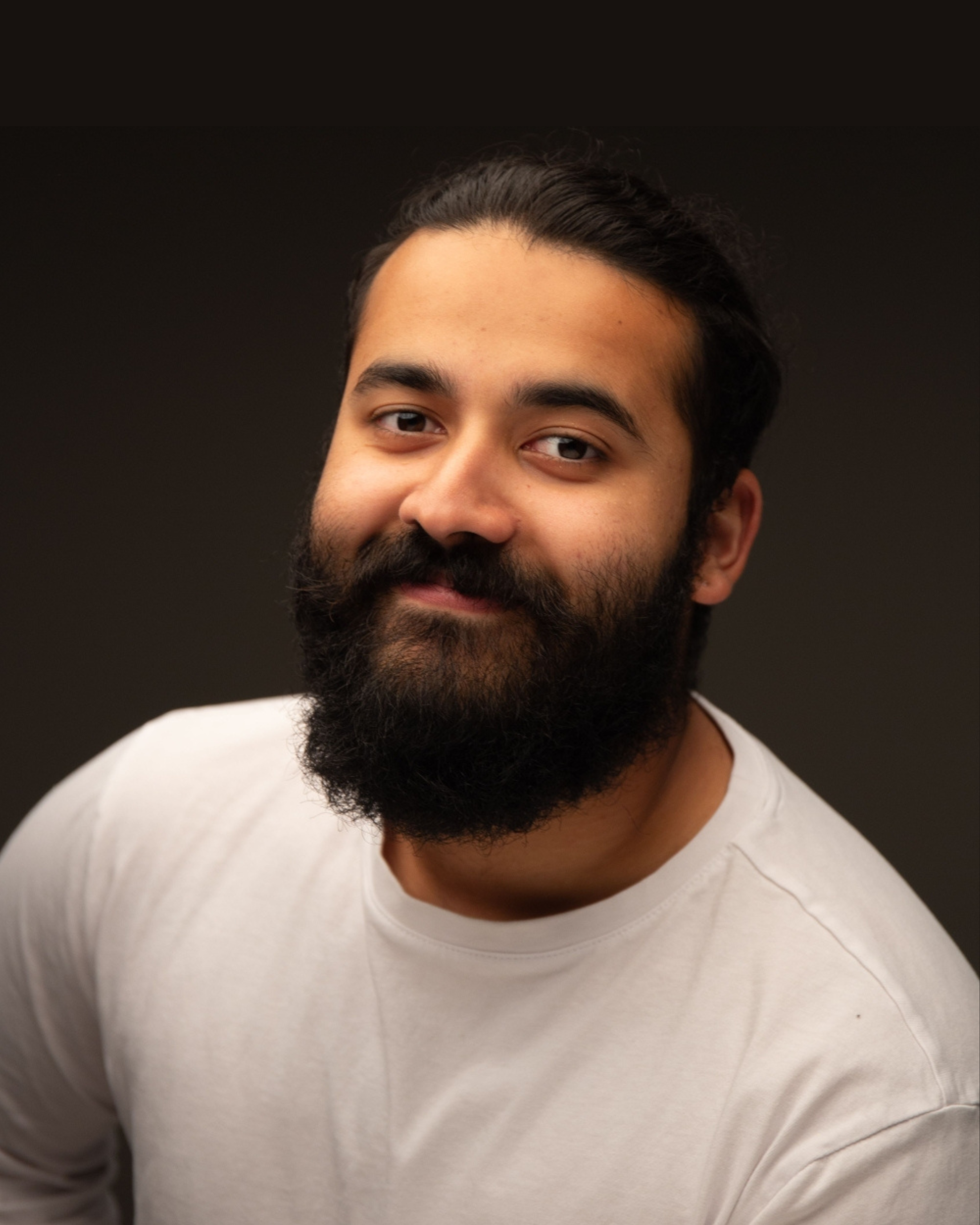 A man with a thick black beard, mustache, and dark hair styled back, wearing a white t-shirt, smiling softly against a dark background.