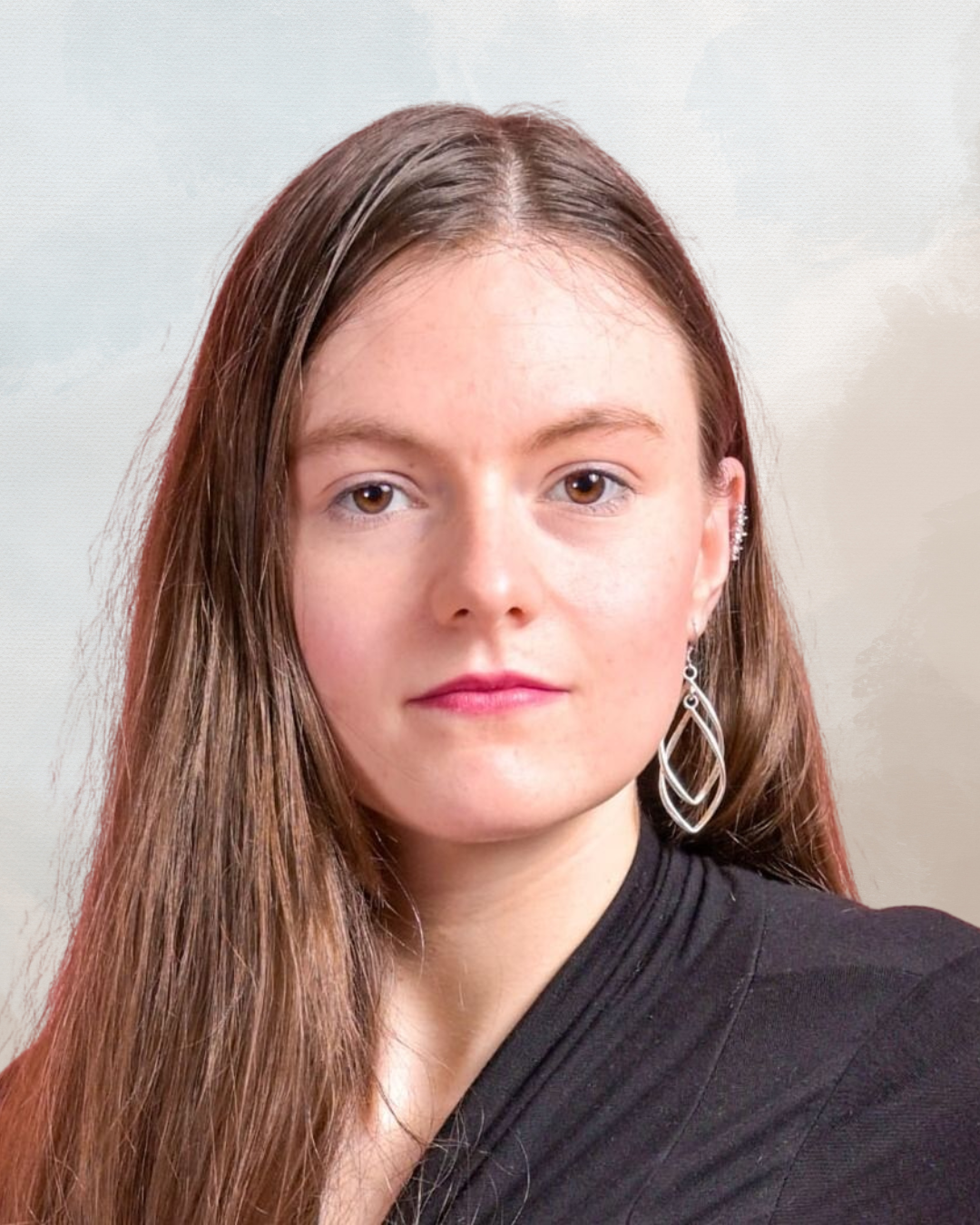 A young woman with long brown hair, brown eyes, and light complexion wearing silver earrings and a black top, looking directly at the camera against a neutral background.